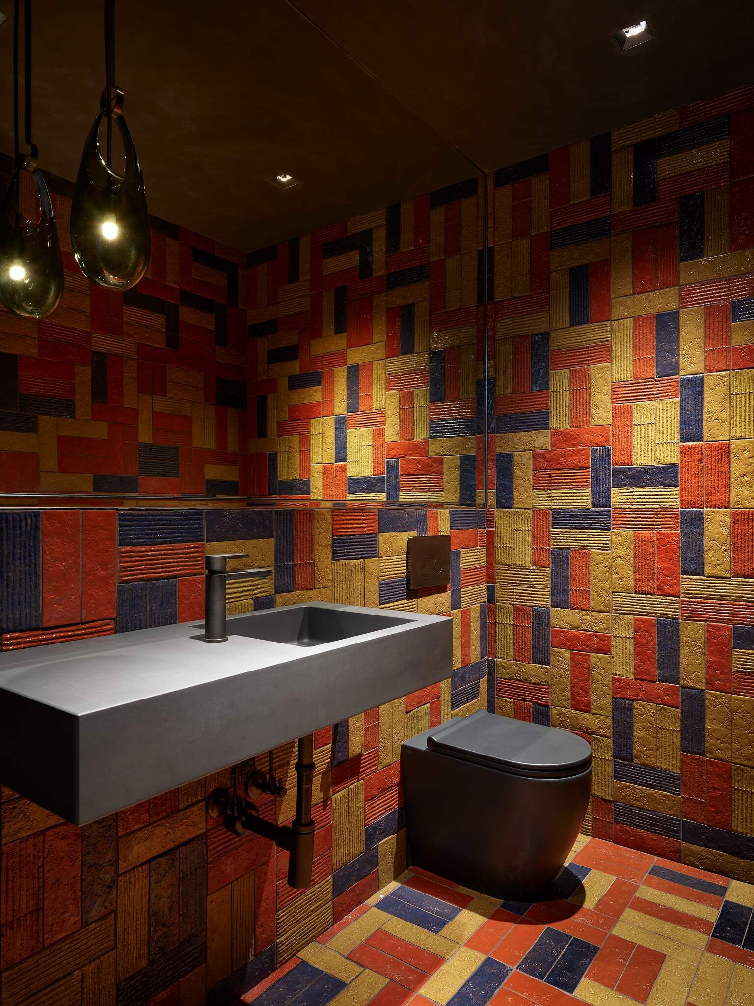 Modern bathroom with vibrant red, gold, and blue geometric patterned tiles covering the walls and floor. Features a wall-mounted gray concrete sink with a matte black faucet, a black toilet, and two distinctive teardrop pendant lights.
