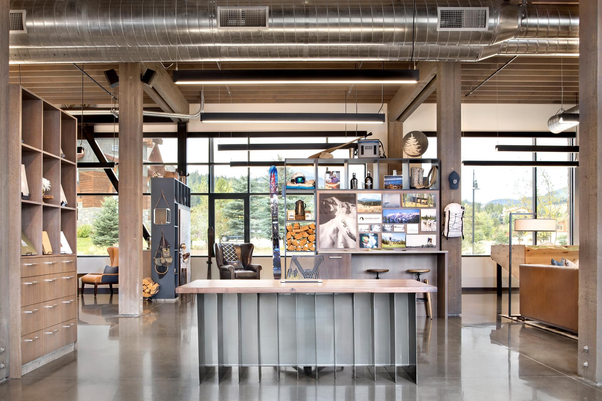 Modern rustic lodge or showroom interior with a long wooden desk and polished concrete floor. A prominent display wall showcases outdoor lifestyle products, ski gear, and mountain landscape photography. Exposed ductwork, wooden beams, and large windows with views of trees and distant mountains complete the contemporary design.