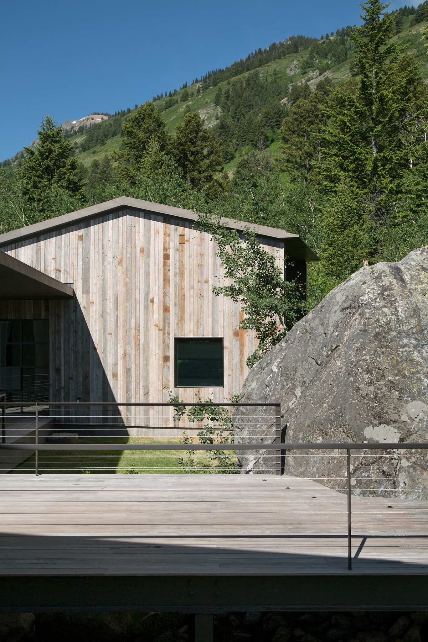 Contemporary mountain home with vertical wood siding and a dark window, featuring a modern wooden deck with cable railings. A large natural rock formation stands beside the house, with a lush, tree-covered mountain and clear blue sky in the background.
