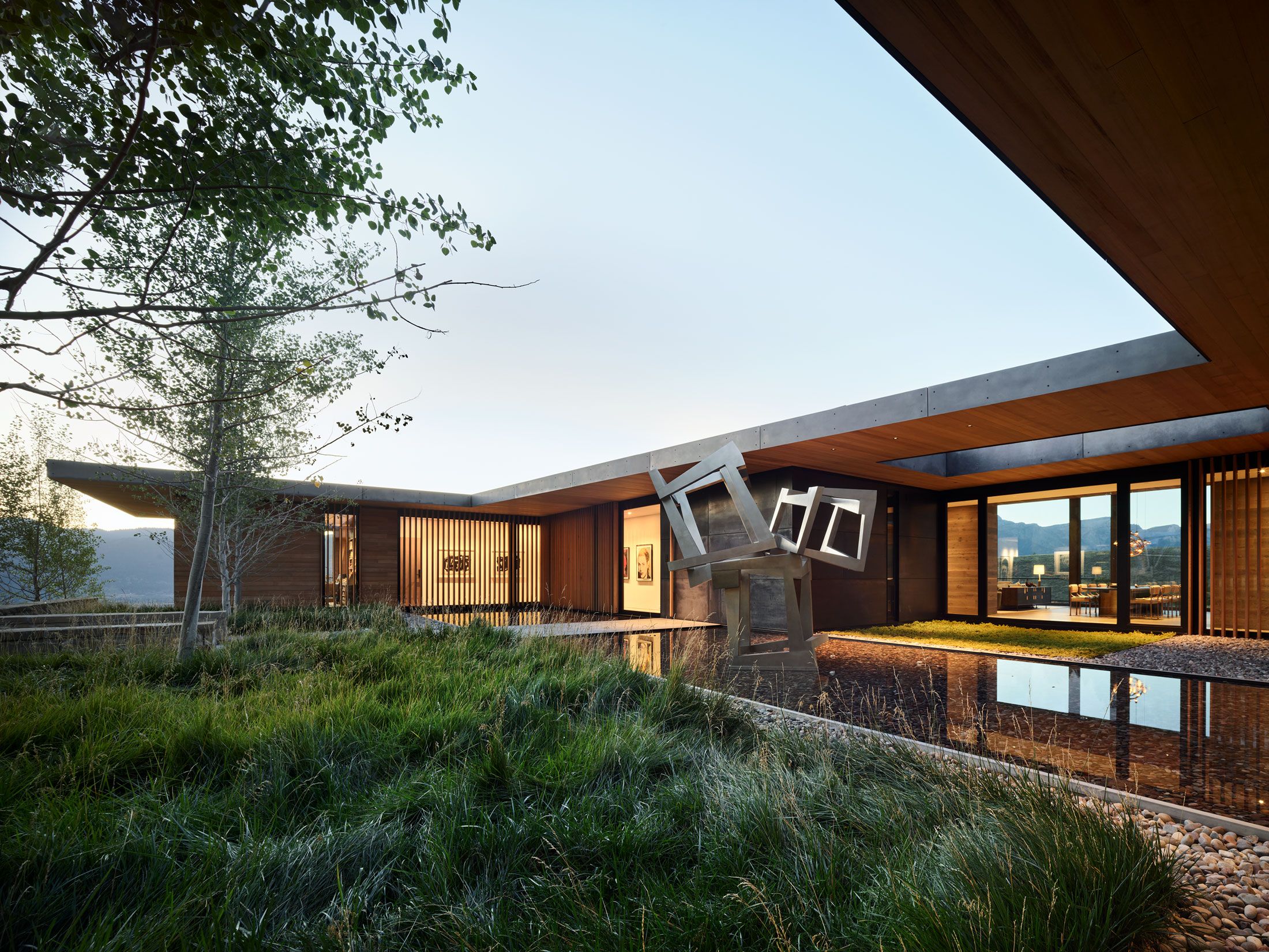 Contemporary luxury home showcasing warm wood and concrete architecture, situated alongside a dark reflective water feature. A striking geometric metal sculpture is prominently placed in the water, bordered by tall natural grass. Illuminated interior spaces are visible through expansive glass windows, set against a bright sky.
