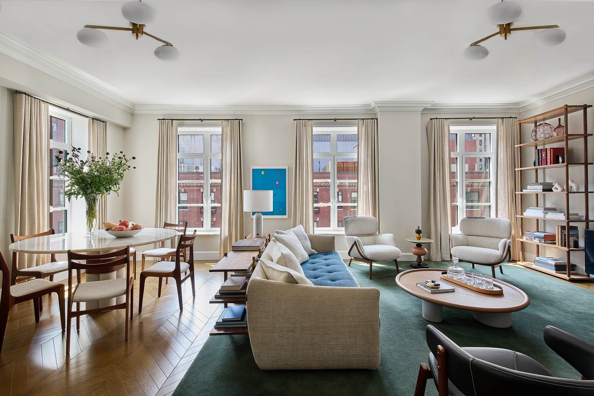 Elegant, open-concept living and dining room featuring a marble oval dining table with mid-century modern wooden chairs, herringbone wood floors, and a bright living area with a contemporary beige and blue sofa, green area rug, and oval coffee table. Large windows with neutral curtains provide ample natural light.