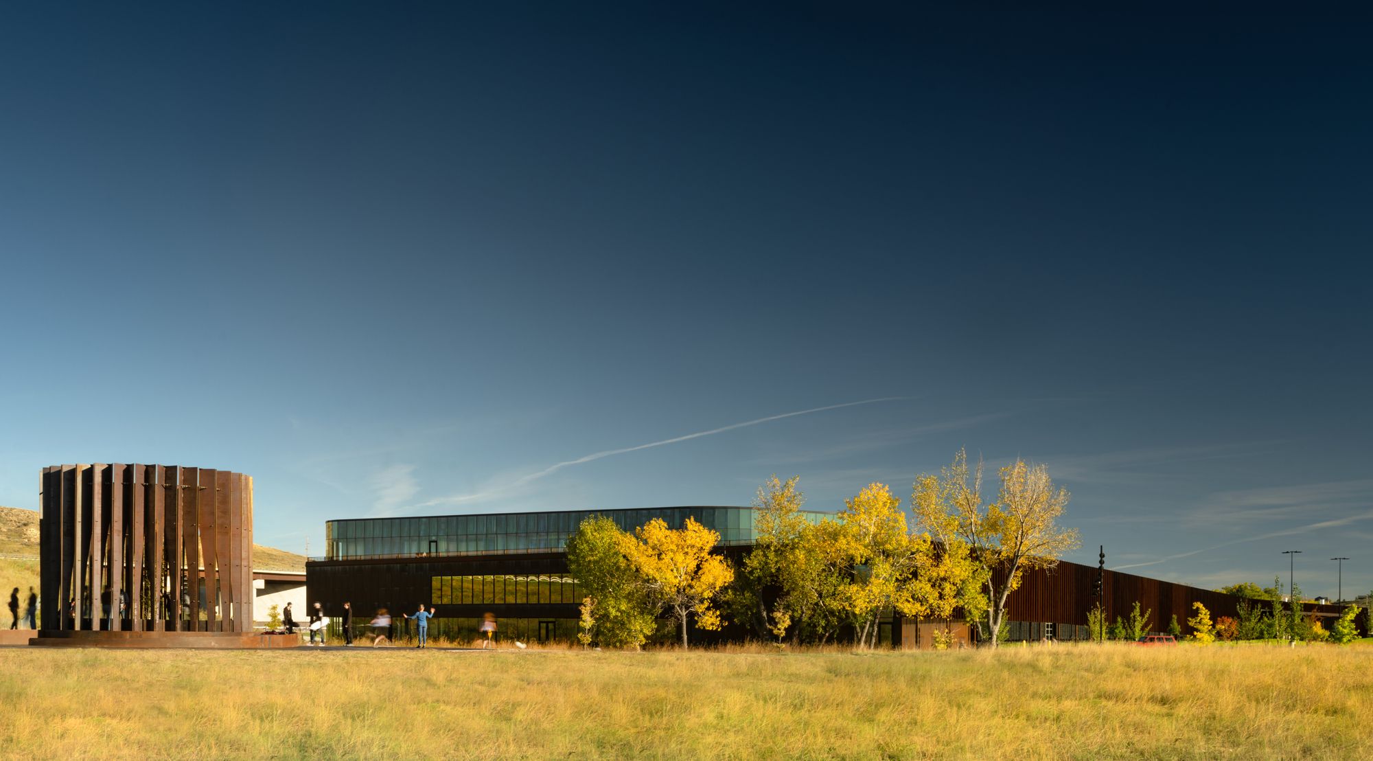 Contemporary architectural complex featuring a circular, weathered Corten steel sculpture and a long building with dark and glass facades. Vibrant yellow autumn trees stand among golden prairie grass in the foreground, with blurred figures moving near the structures under a deep blue sky.