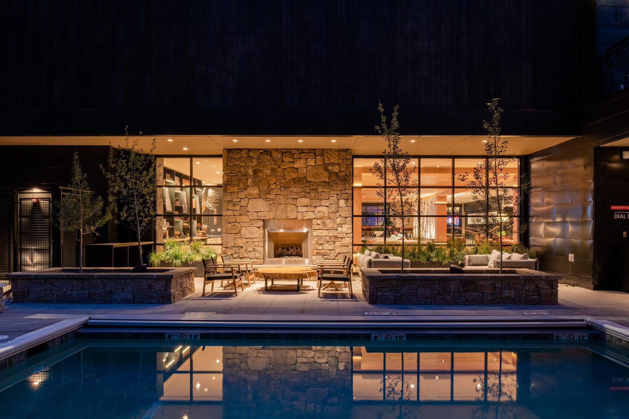 Nighttime view of an illuminated outdoor pool and patio area. The foreground shows a swimming pool reflecting warm lights from the adjacent building. On the paved patio, there's a prominent stone-clad outdoor fireplace, comfortable lounge seating, and wooden tables. Small trees in stone planters are placed between the seating and the modern building's large windows, which reveal brightly lit interior spaces.