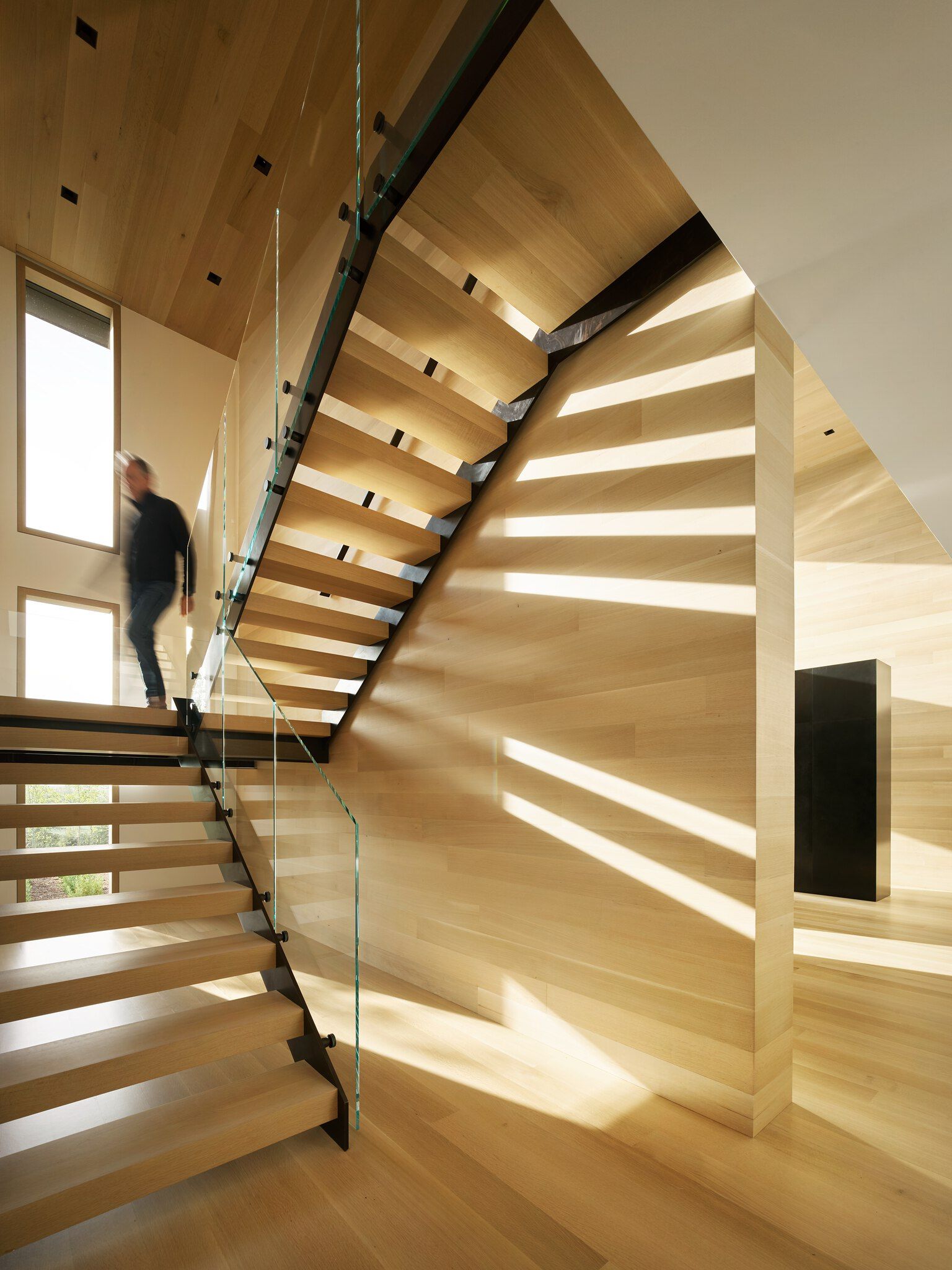 Modern architectural interior featuring a sleek light wood staircase with a glass balustrade and black metal supports. A blurred figure descends the stairs as dramatic sunlight creates striking parallel shadows across the adjacent wood-paneled wall and steps. The space is filled with warm light wood surfaces on floors, walls, and a vaulted ceiling, with a dark rectangular accent visible in the background.