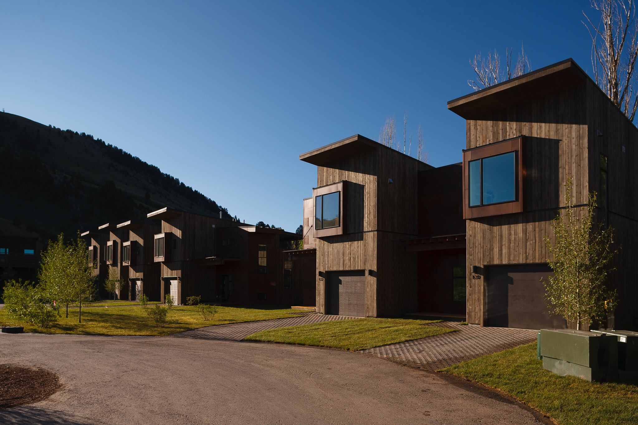 Modern wooden homes with large windows and overhanging roofs line a paved road with green lawns, backed by a shadowed mountain under a clear blue sky.