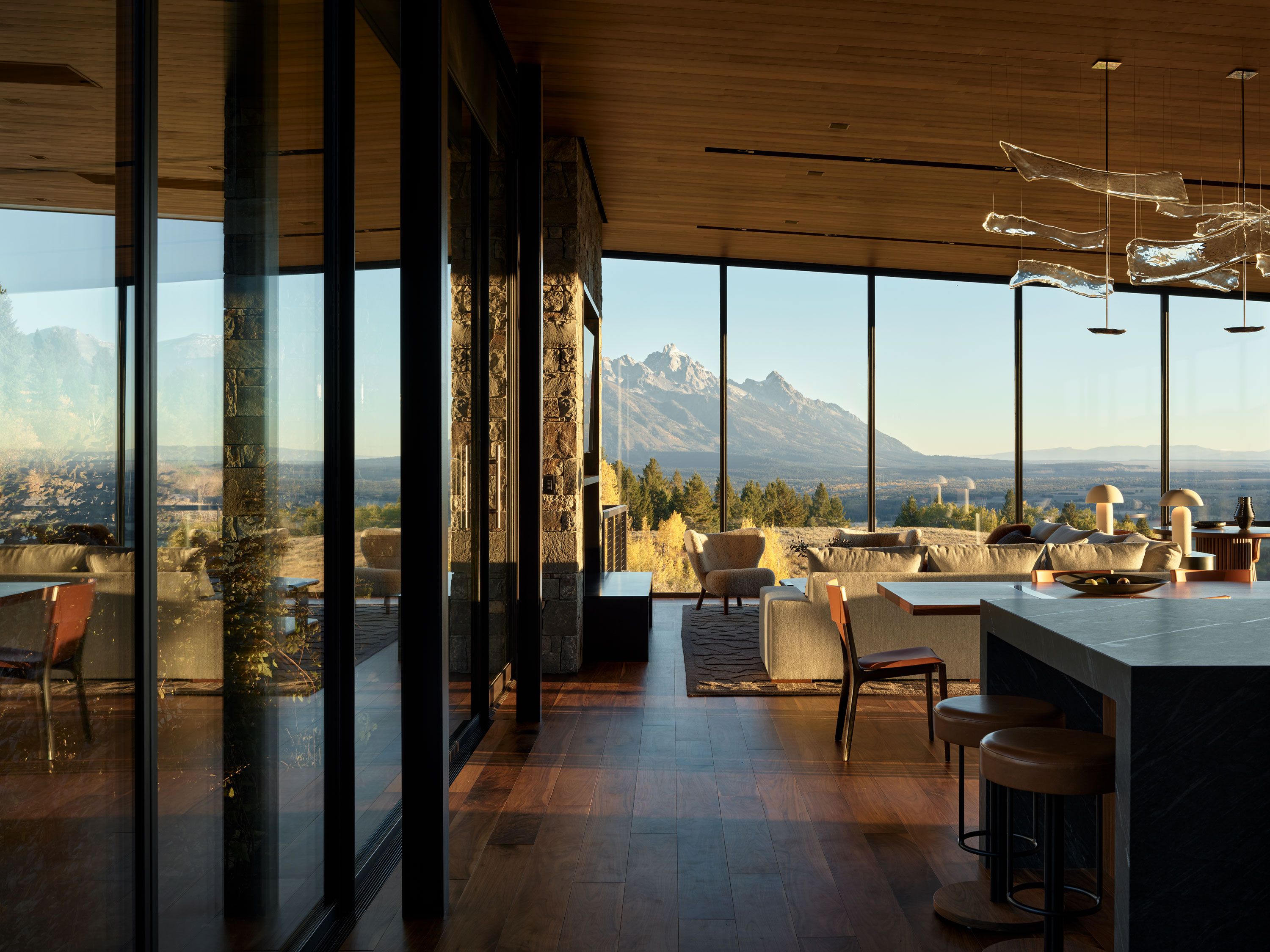 Modern luxury home interior with open concept living and kitchen, featuring floor-to-ceiling windows overlooking a panoramic view of snow-capped mountains. The space includes warm wood ceilings, dark wood floors, and a stone fireplace pillar.