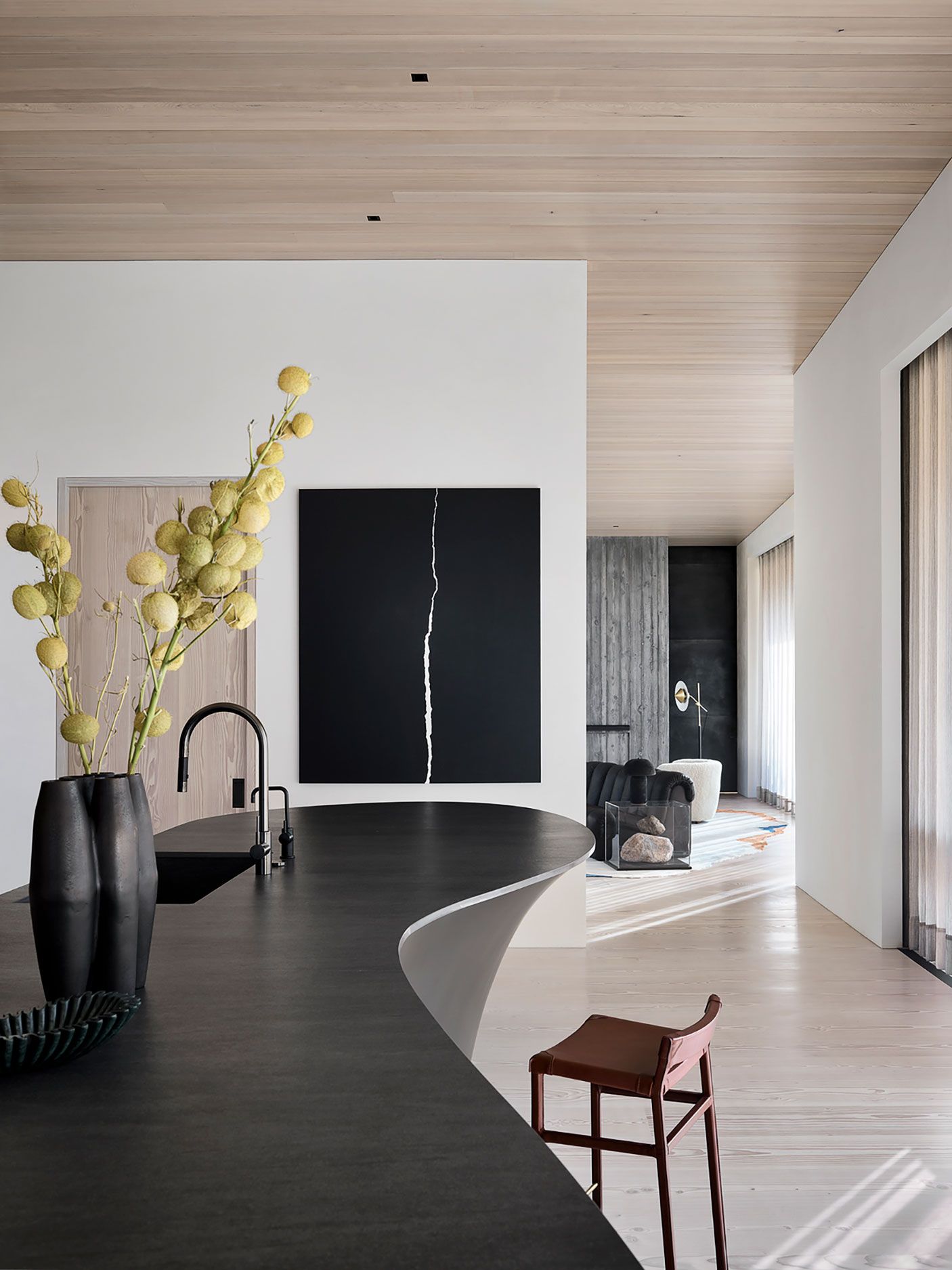 Minimalist kitchen featuring a curved dark island, wooden ceilings, light floorboards, and a large black art piece.