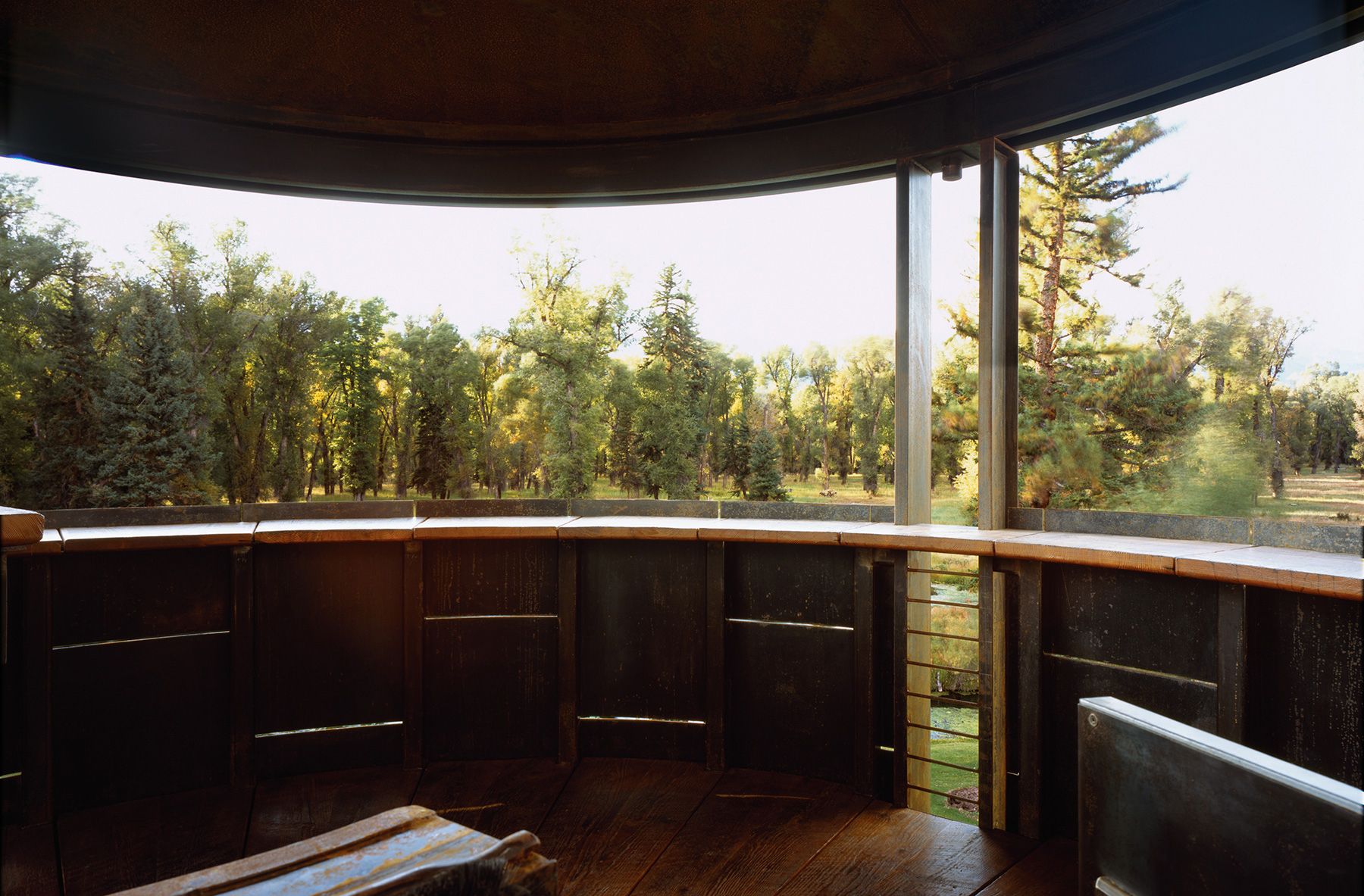 Interior view from an elevated circular observation deck featuring dark metal walls, wooden flooring, and wide open-air windows overlooking a lush forest.