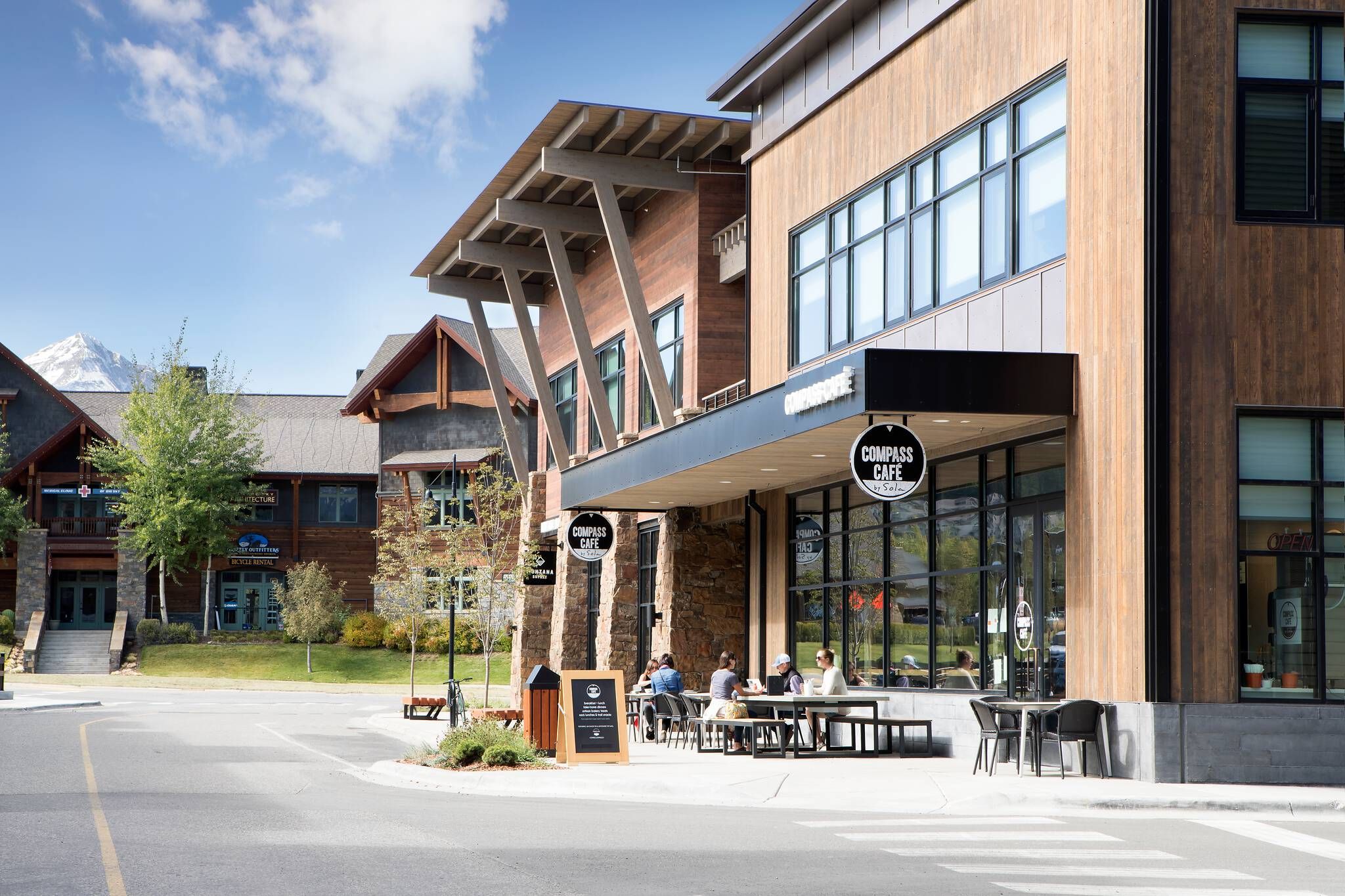 Compass Cafe with outdoor dining and patrons, set in a modern mountain village street with contemporary wood and stone buildings, and a snow-capped mountain in the background under a blue sky.
