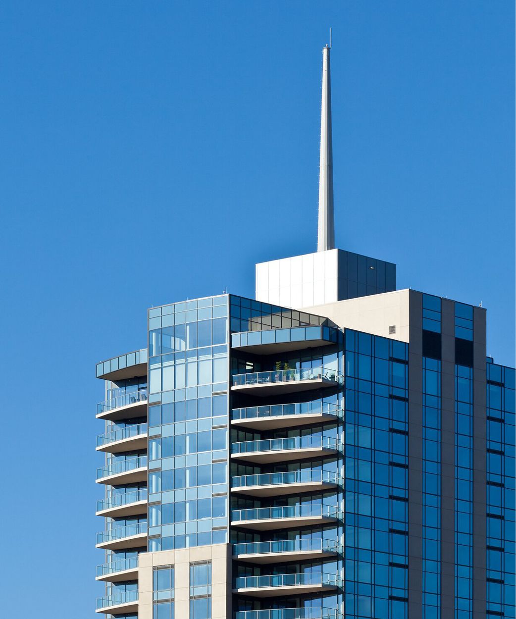 Modern high-rise building with a blue reflective glass facade, multiple balconies with glass railings, and a slender white spire against a clear blue sky.
