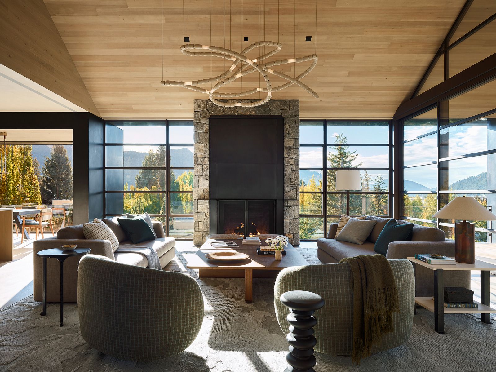 Luxury living room interior featuring a large stone fireplace, plush sofas, modern light fixture, and floor-to-ceiling windows with mountain views.
