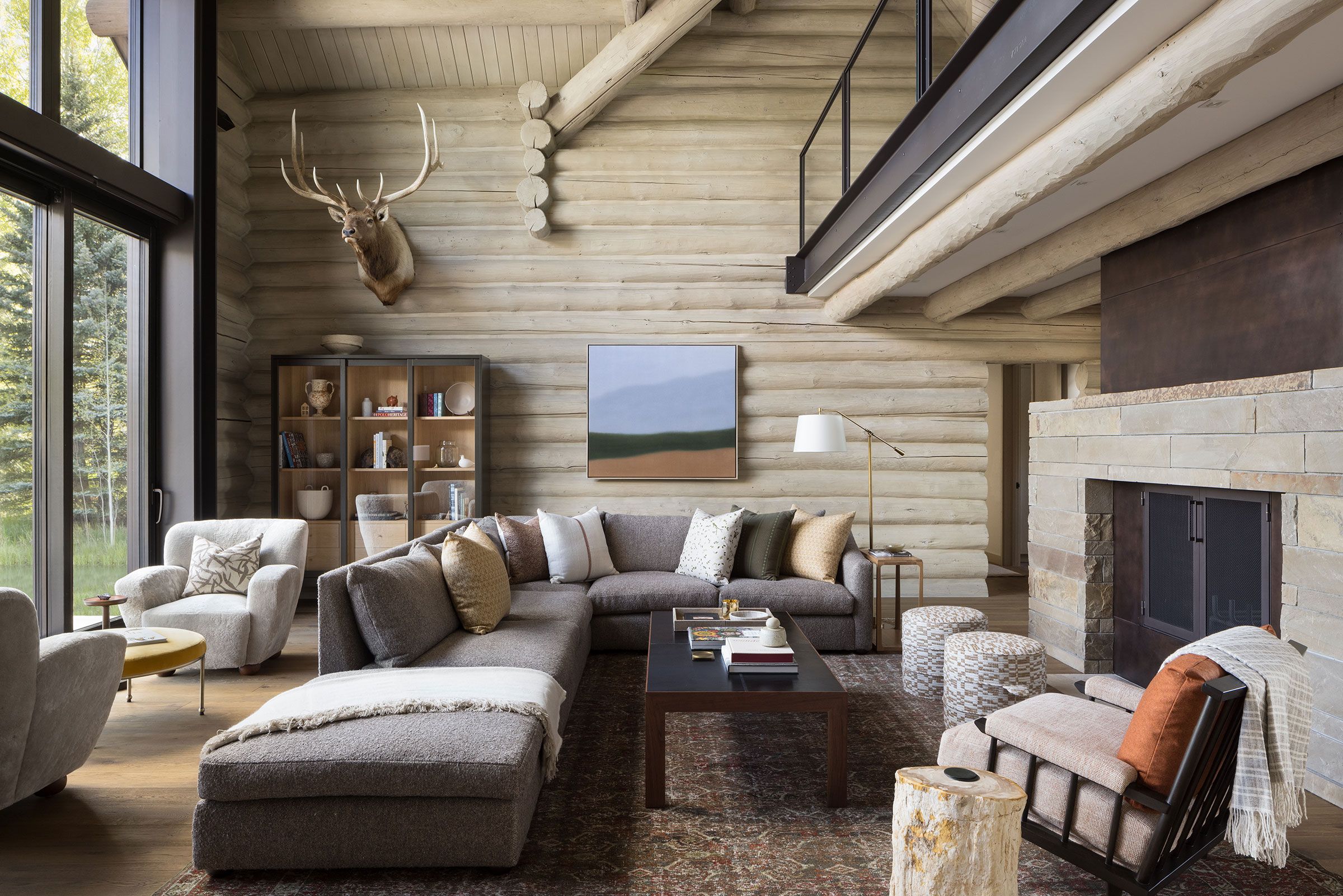 Rustic modern living room with a large gray sectional sofa, stone fireplace, and exposed log walls under a high ceiling.