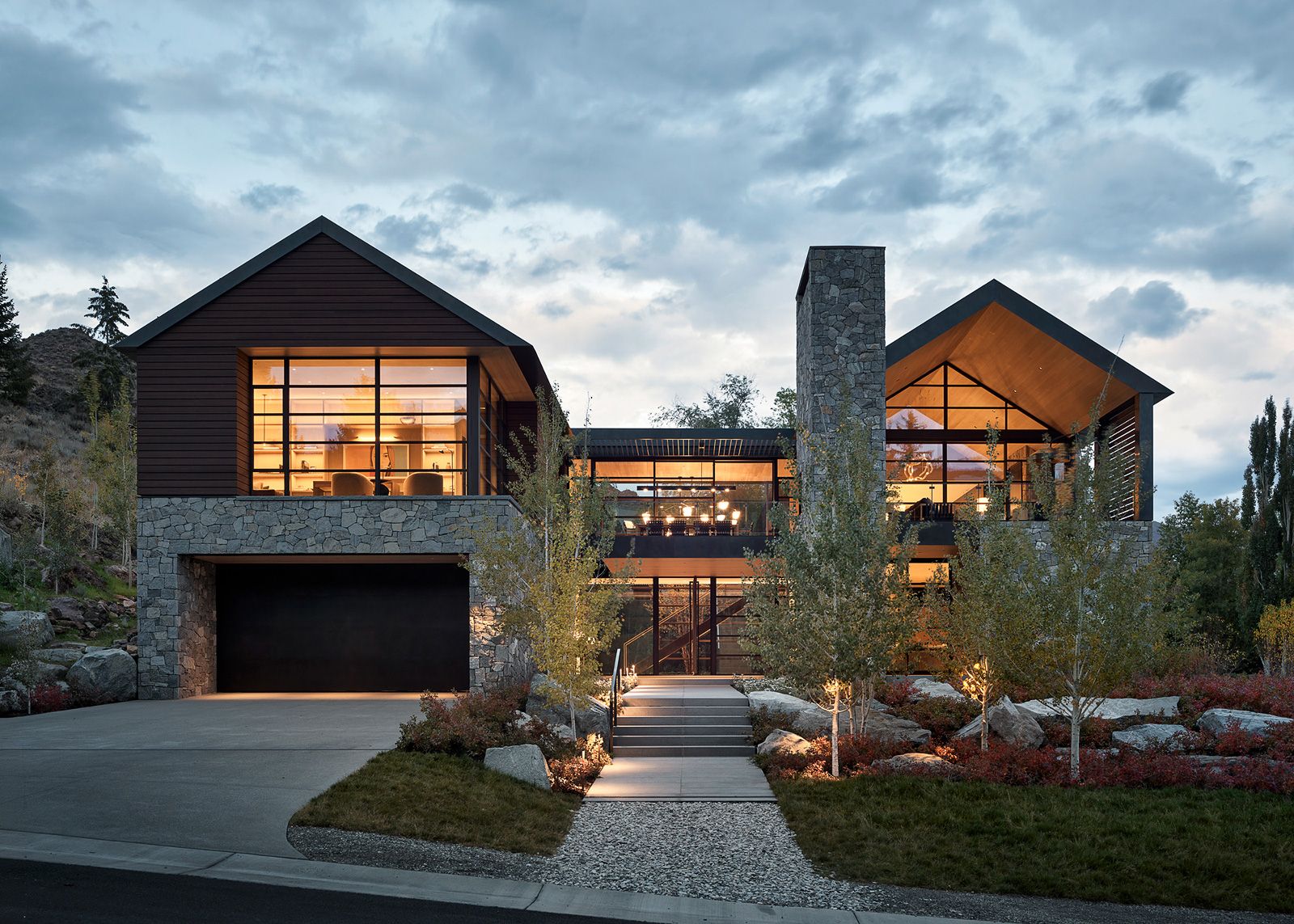 Elevated view of a luxury mountain home with glowing windows overlooking a valley town at dusk under a cloudy sky.