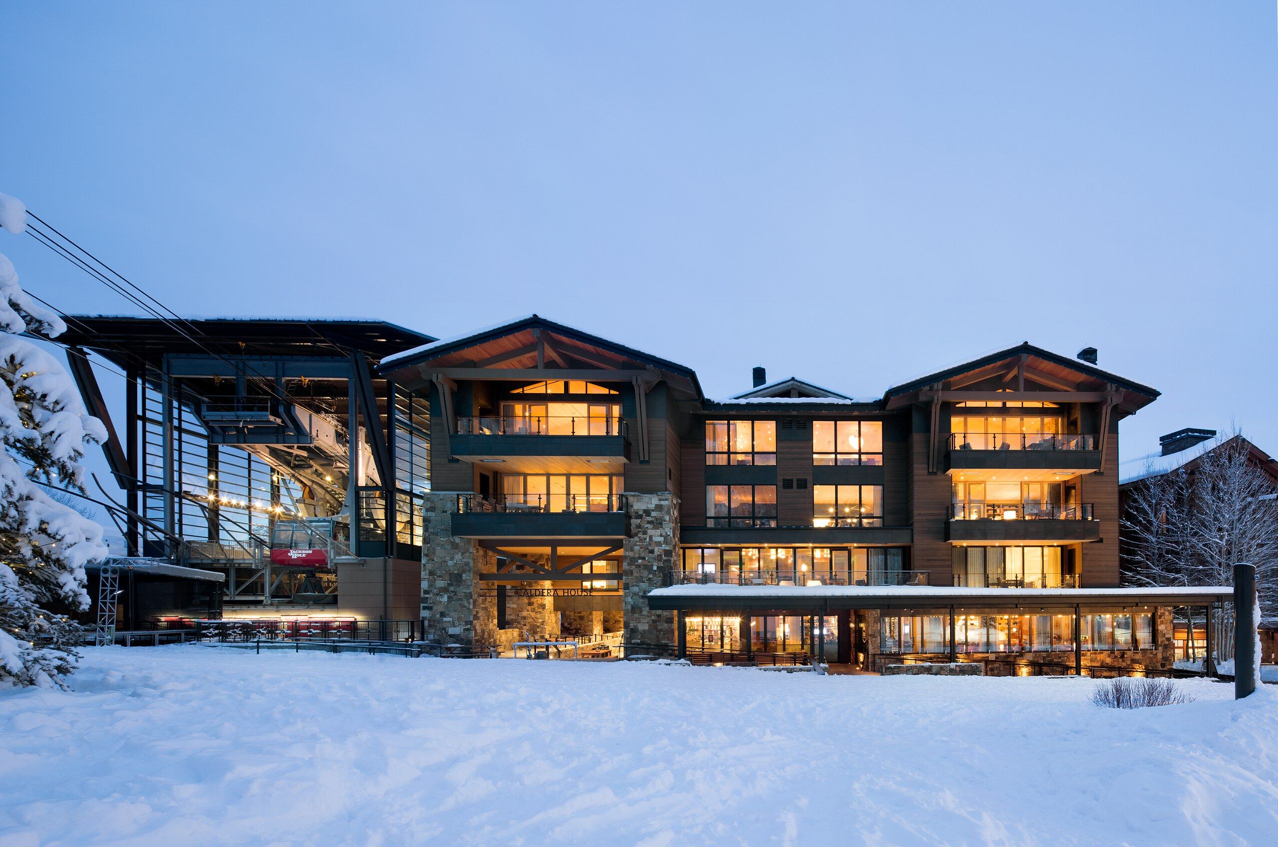 Caldera House luxury ski lodge in Jackson Hole, Wyoming, at dusk. The multi-story building with glowing windows, balconies, and stone accents stands next to a ski gondola station, all surrounded by deep snow under a twilight sky.