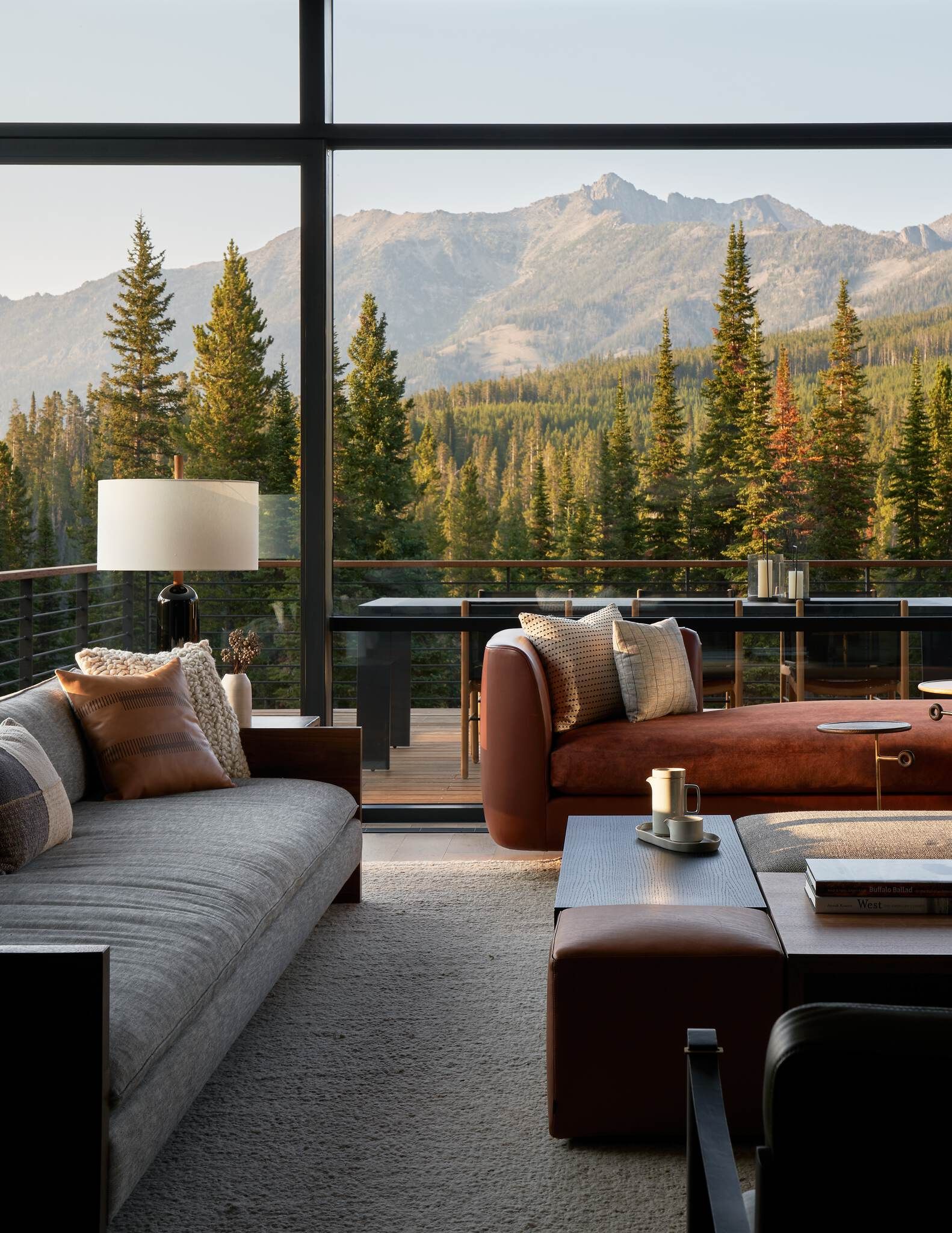 Modern living room with expansive windows showcasing a majestic mountain range and dense pine forest. A gray fabric sofa, rust-toned chaise lounge, and dark wood coffee tables are arranged on a light textured rug, with an outdoor deck visible.