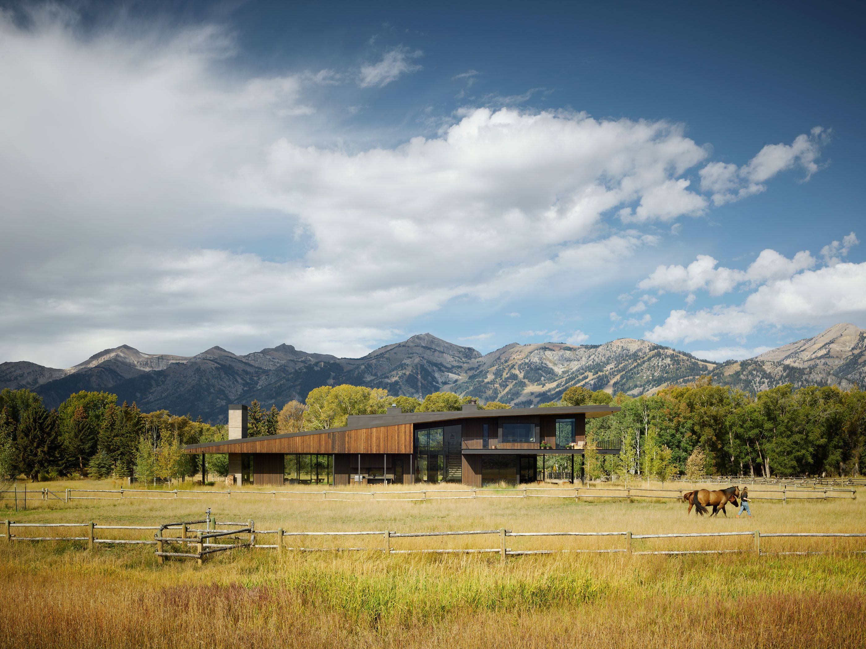 A modern wooden and glass house nestled in a mountainous landscape with vibrant fall foliage. A golden field with a wooden fence extends to the foreground, where a person leads two horses.