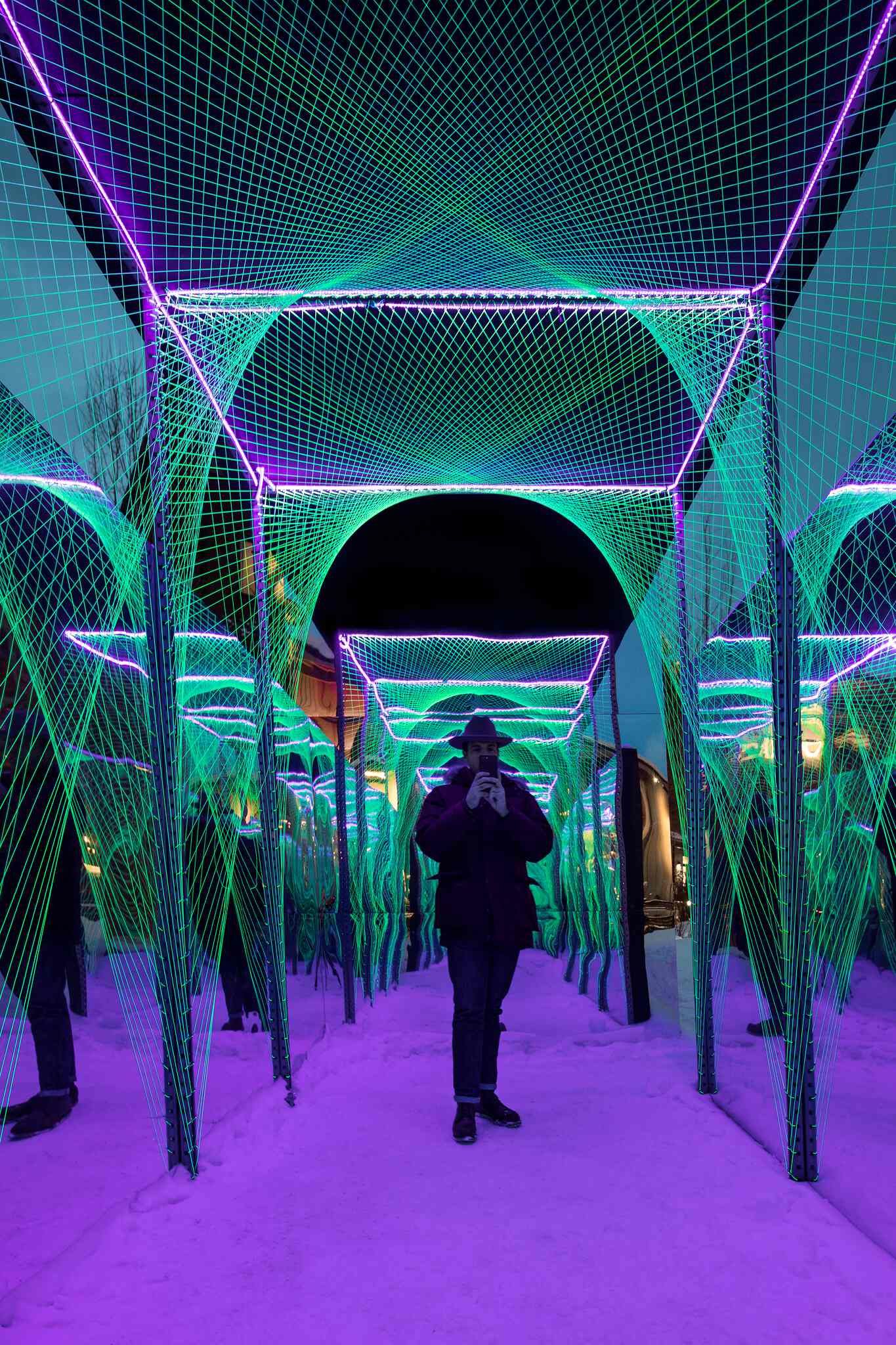 Person in a dark jacket and hat holding a smartphone, standing on snow inside an immersive light art installation. The installation features neon green grid-like structures and arches, illuminated by vibrant purple lights that also cast a glow on the snowy ground.