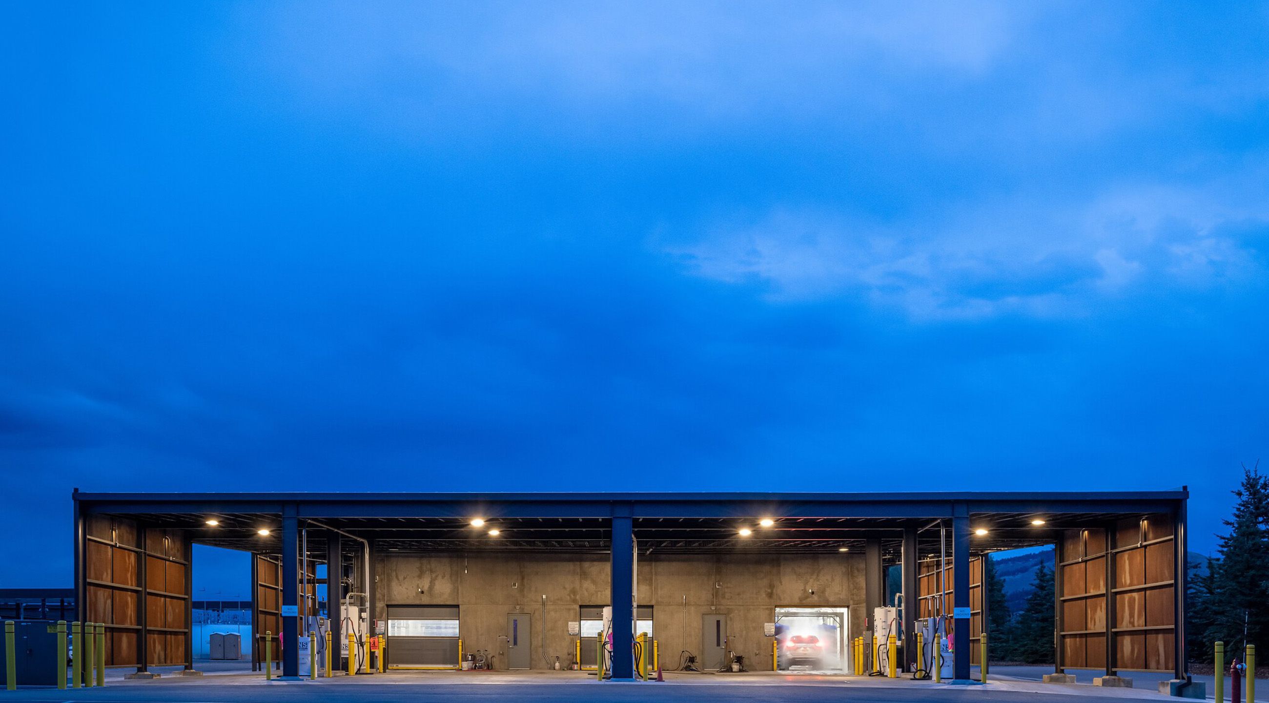 Modern car wash facility with several illuminated bays at dusk, featuring a car being washed inside one bay. The scene is set under a deep blue sky with distant mountains and evergreen trees.