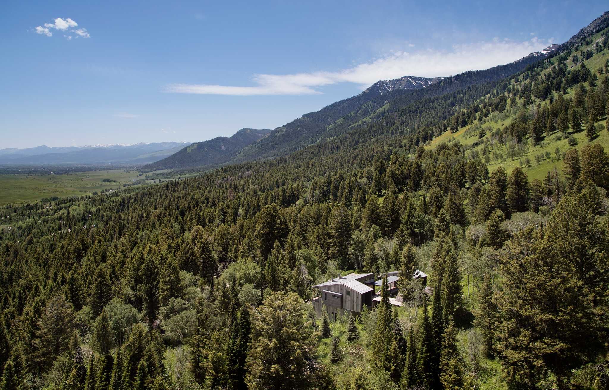 Aerial view of a contemporary house with a multi-pitched roof, nestled in a dense evergreen forest. An expansive green valley stretches into the distance, framed by forested mountains and distant snow-capped peaks under a clear blue sky.