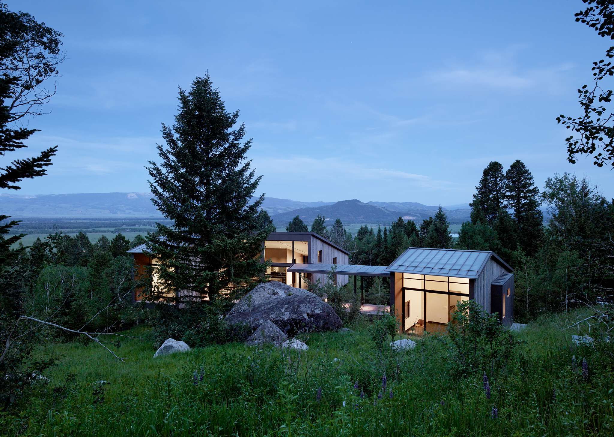A modern architectural home, featuring multiple structures with wood and metal siding, large illuminated windows, and a connecting walkway, nestled on a lush, boulder-strewn hillside amidst tall evergreen trees, with a scenic valley and distant mountains under a twilight sky.