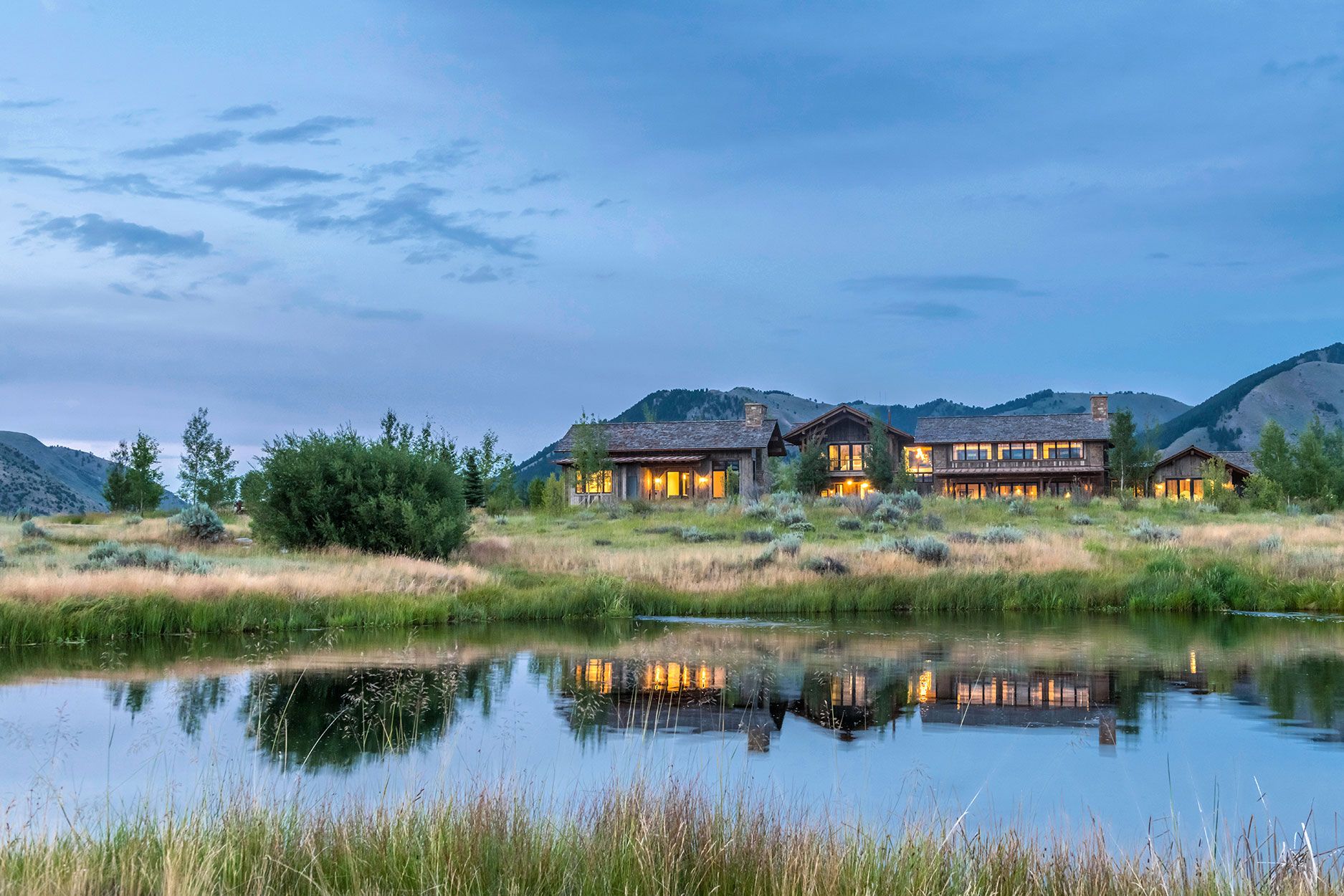 An exterior twilight view of a luxury rustic stone and wood home illuminated by warm lights, sitting on a grassy hill with its reflection cast across a calm pond in the foreground.
