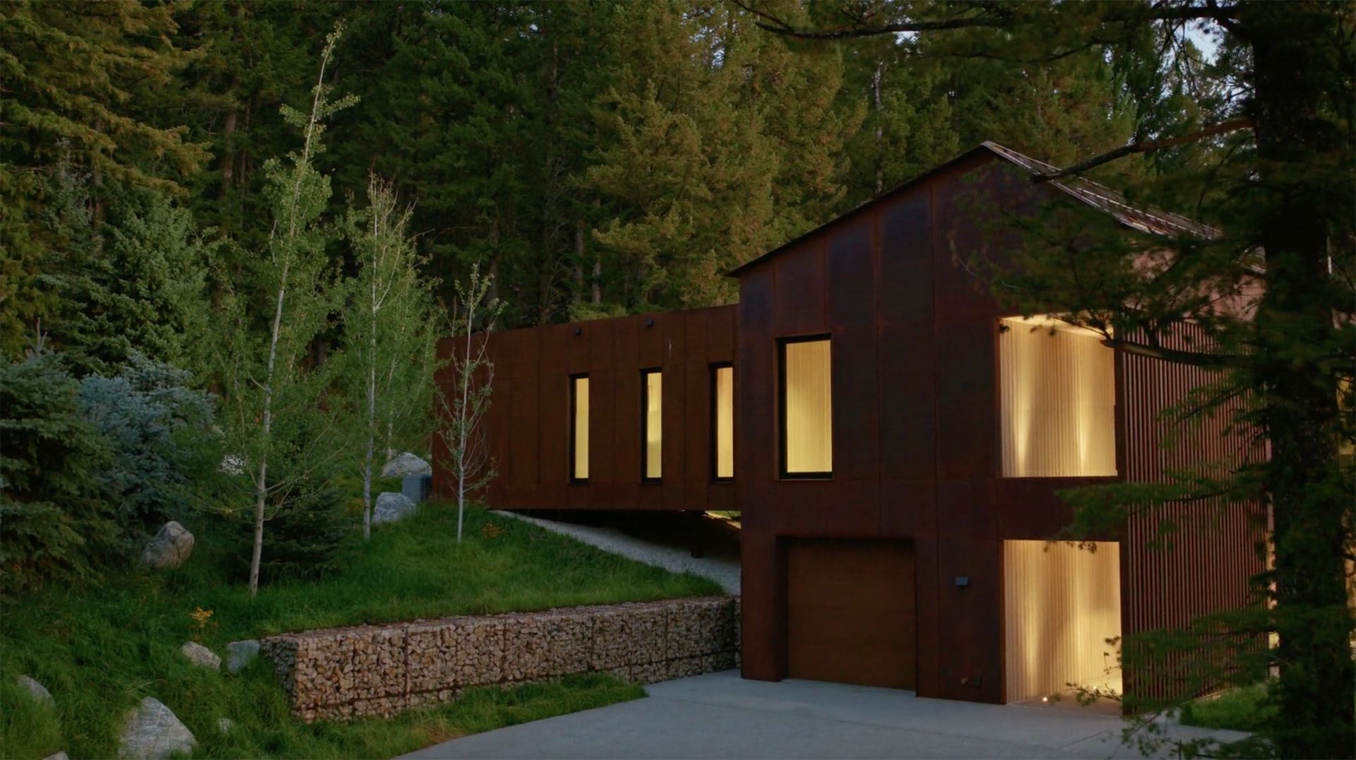Contemporary house with a dark, rusted metal exterior nestled in a dense forest, featuring a cantilevered section, illuminated windows, and a gabion retaining wall.