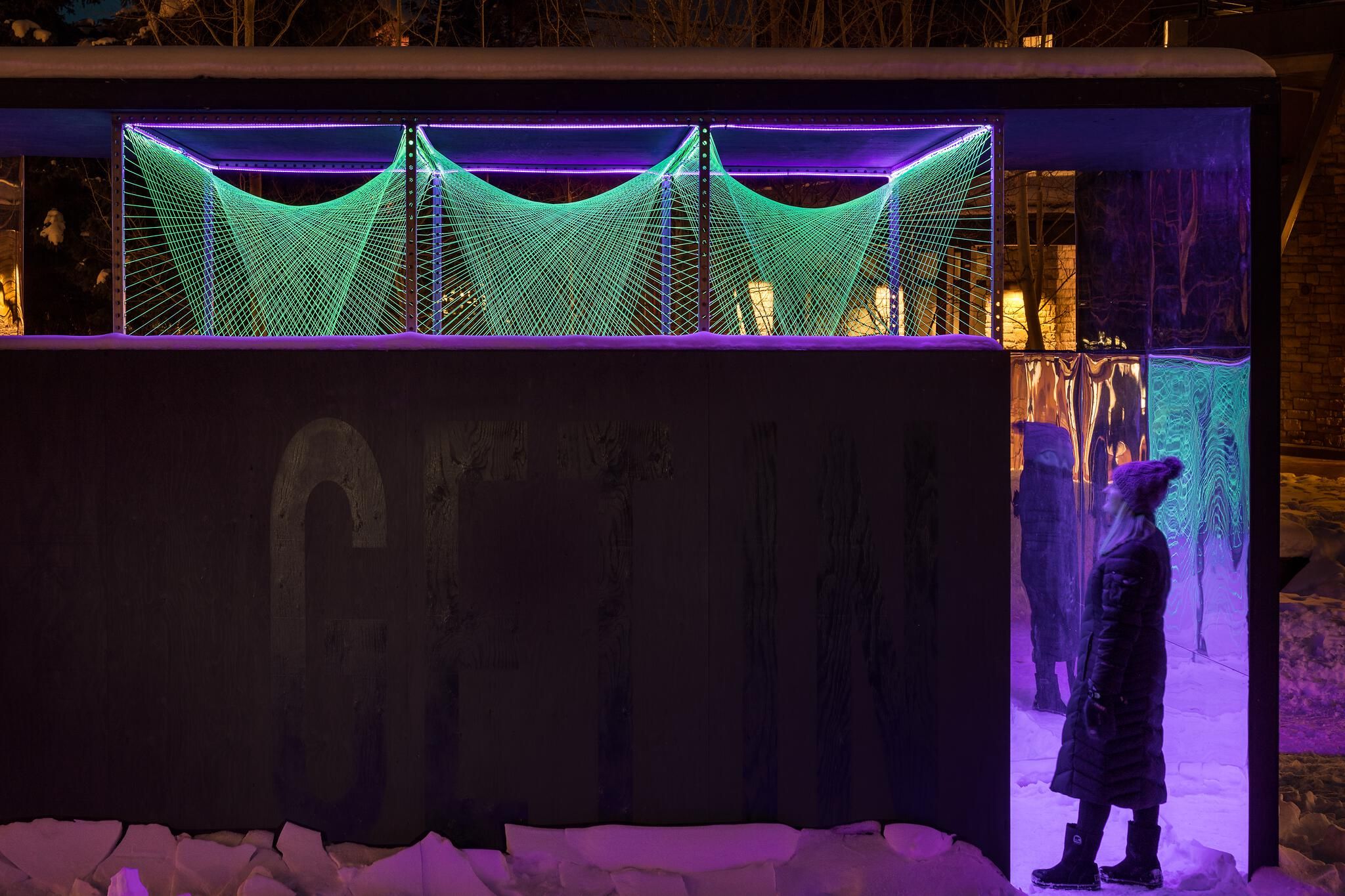 Nighttime outdoor art installation with a dark structure featuring glowing green strings forming intricate geometric patterns. Purple LED lighting illuminates the upper frame and snow-covered ground. A woman in a winter coat observes the reflective panels and light display.