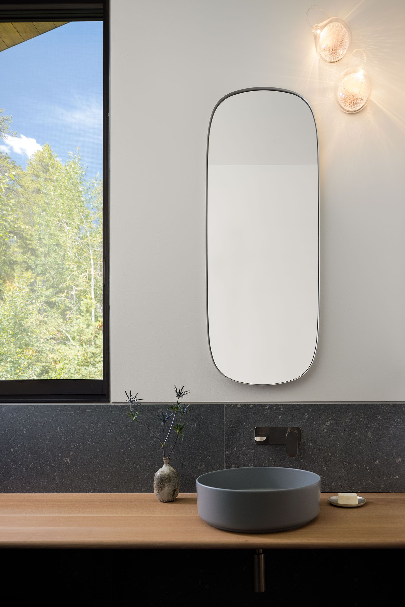 Minimalist bathroom vanity featuring a round gray vessel sink on a light wood countertop, a tall oval mirror, and a dark slate backsplash beneath a tall window.