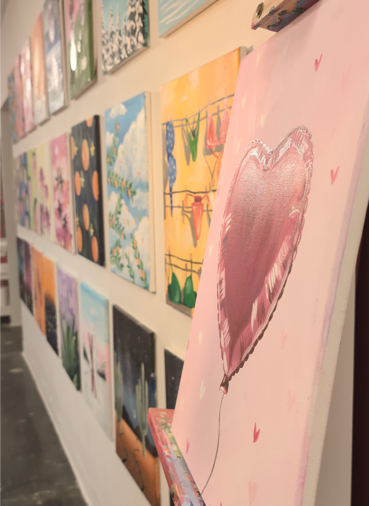 A gallery wall of Art from the Heart's paint night options. Pictured are a lemon and orange painting on a blue striped background, and watermelon on a green background.