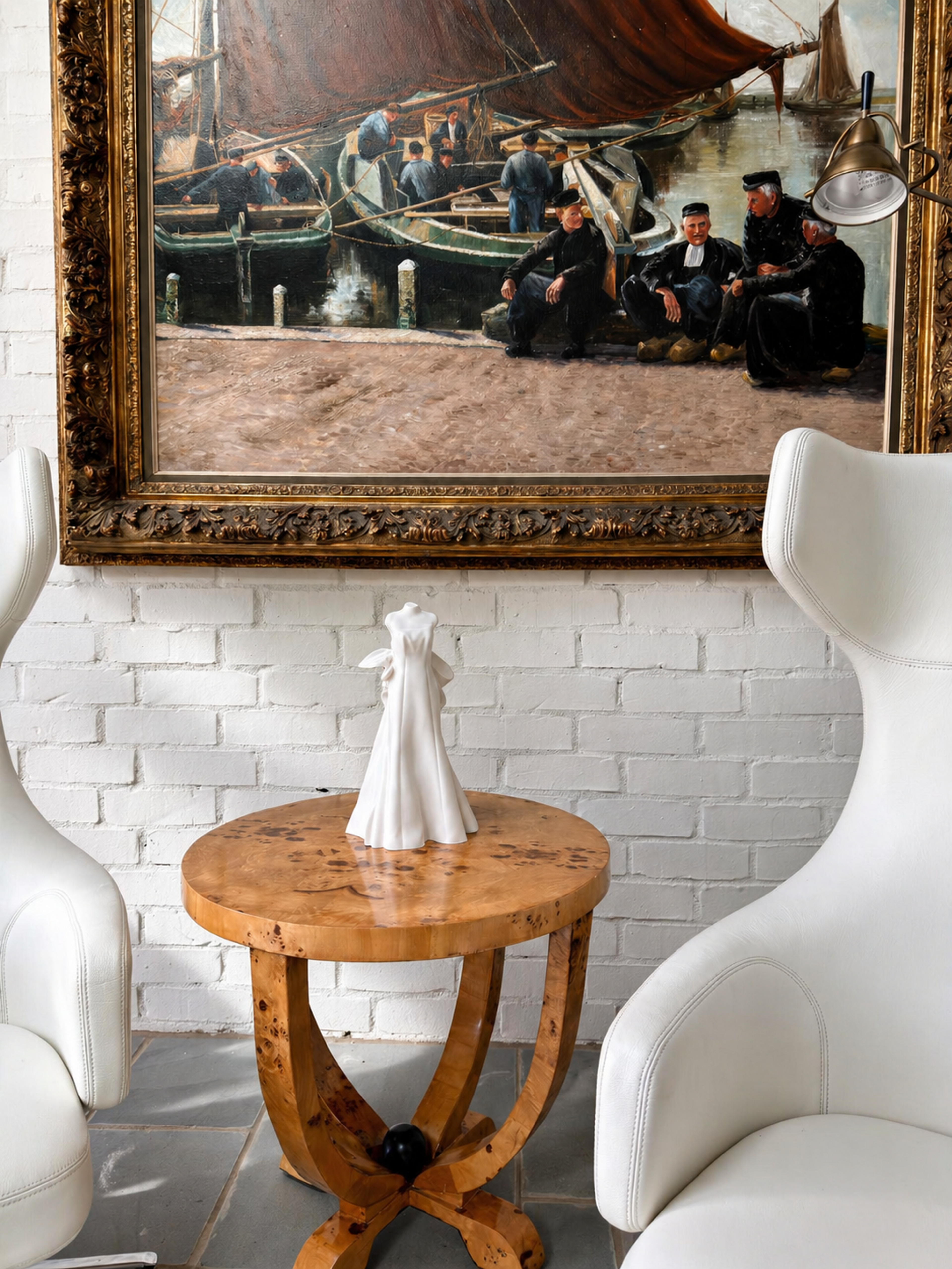 White wedding gown sculpture displayed on a round wooden table between two white lounge chairs beneath a framed painting in a residential interior.