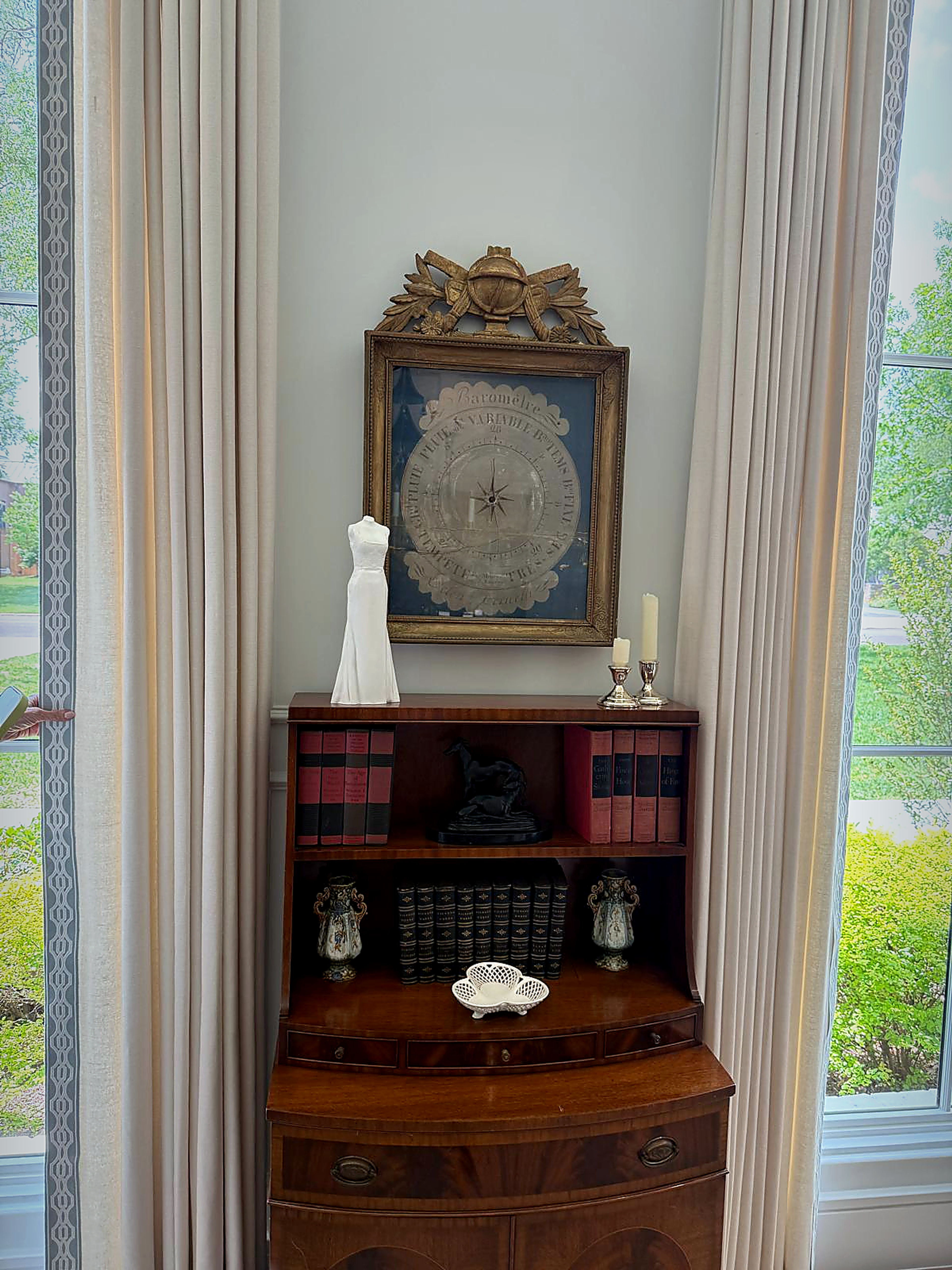 White wedding gown sculpture displayed on an antique wooden cabinet between tall windows beneath a framed artwork in a residential interior.