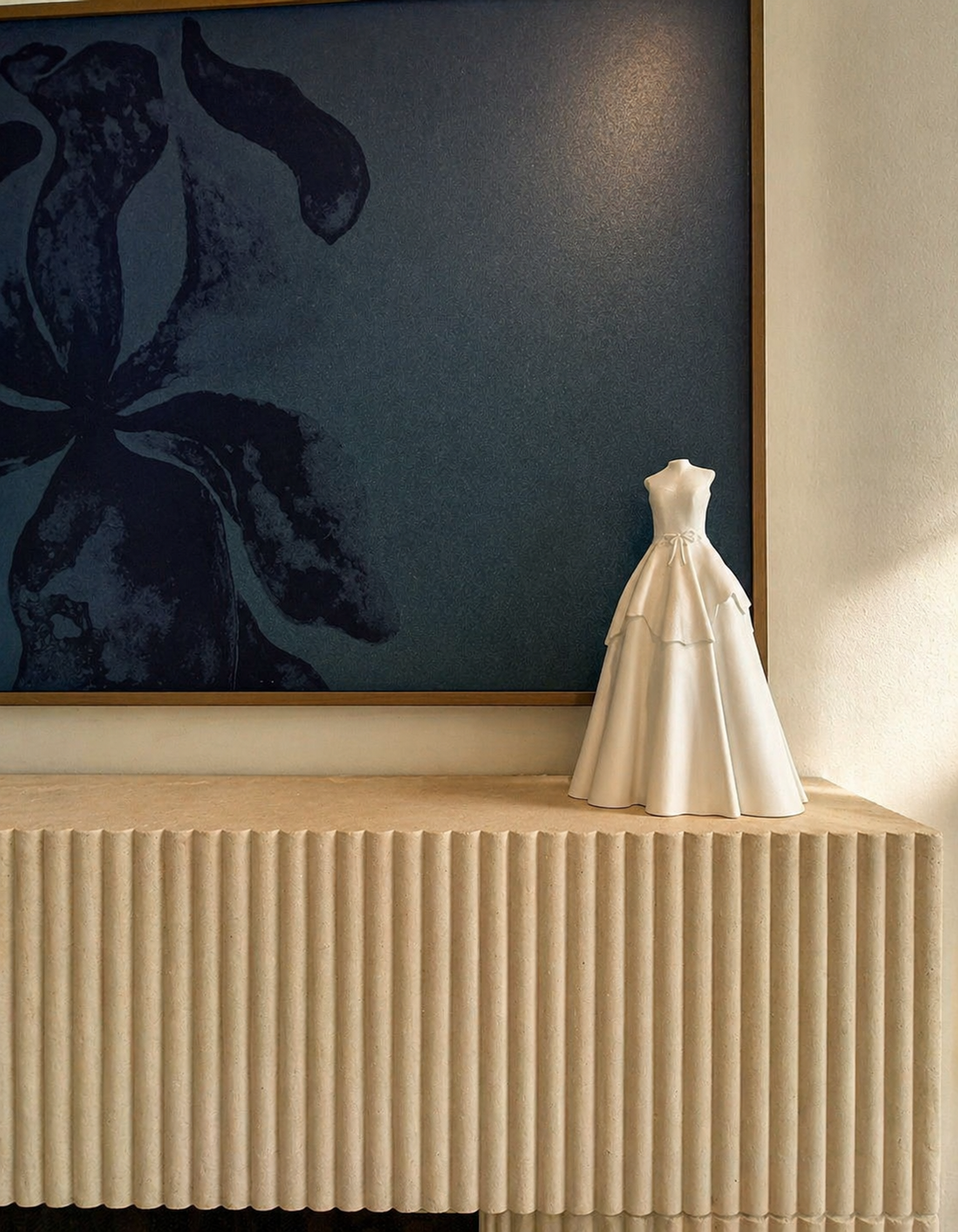 White wedding gown sculpture displayed on a fluted cabinet beneath a large framed artwork.