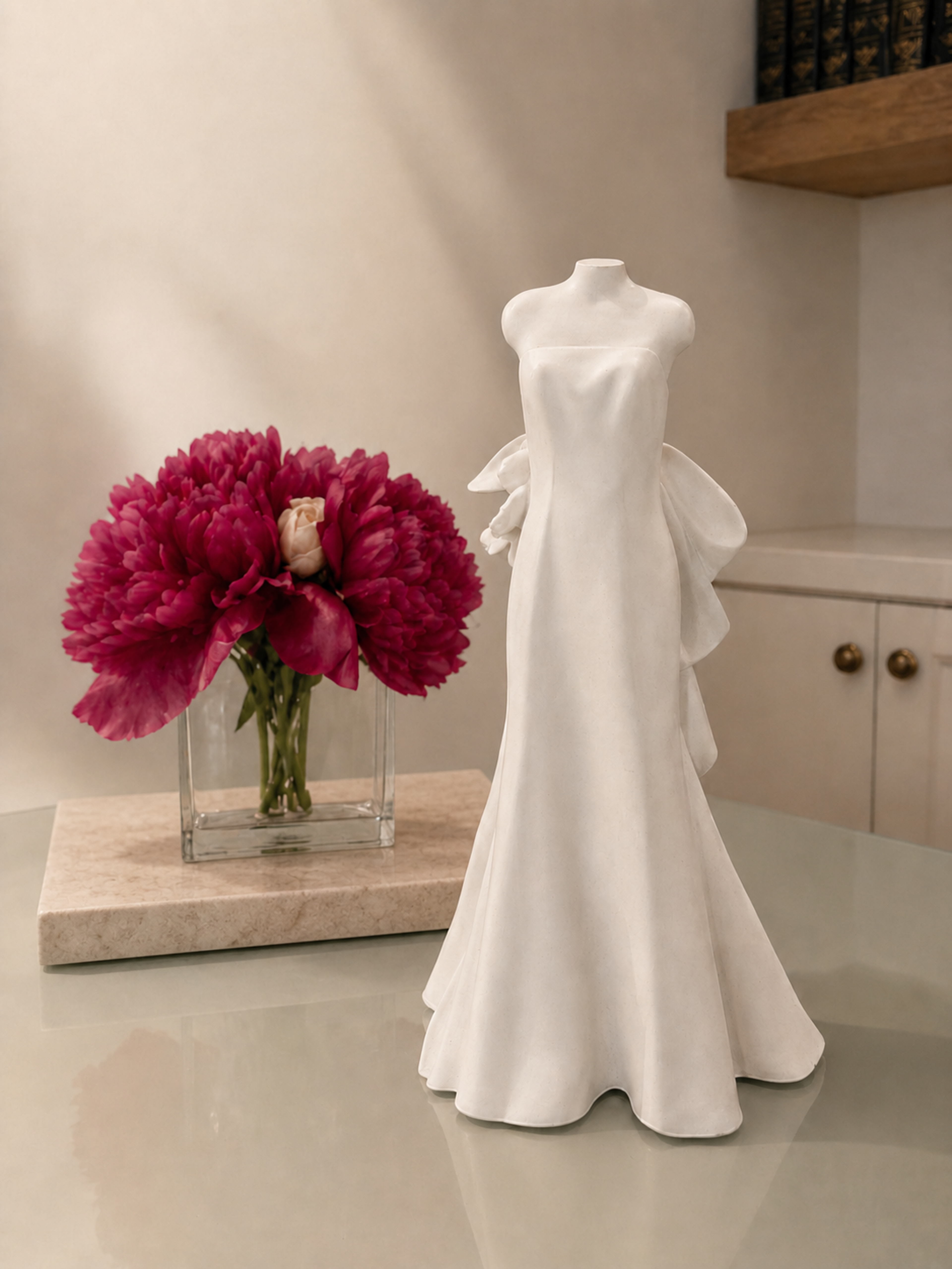 White wedding gown sculpture displayed beside a vase of pink peonies on a stone countertop in a residential interior.