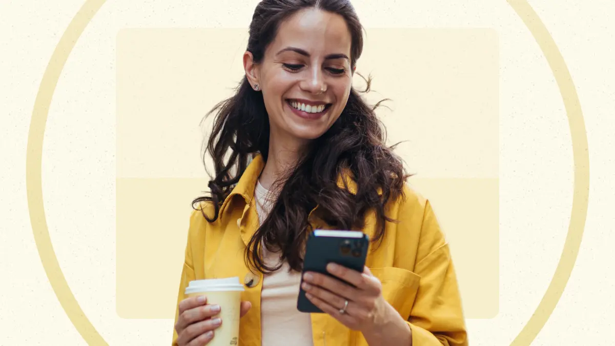 A woman in a yellow jacket holds a coffee cup and smiles while looking at her smartphone against a beige background.