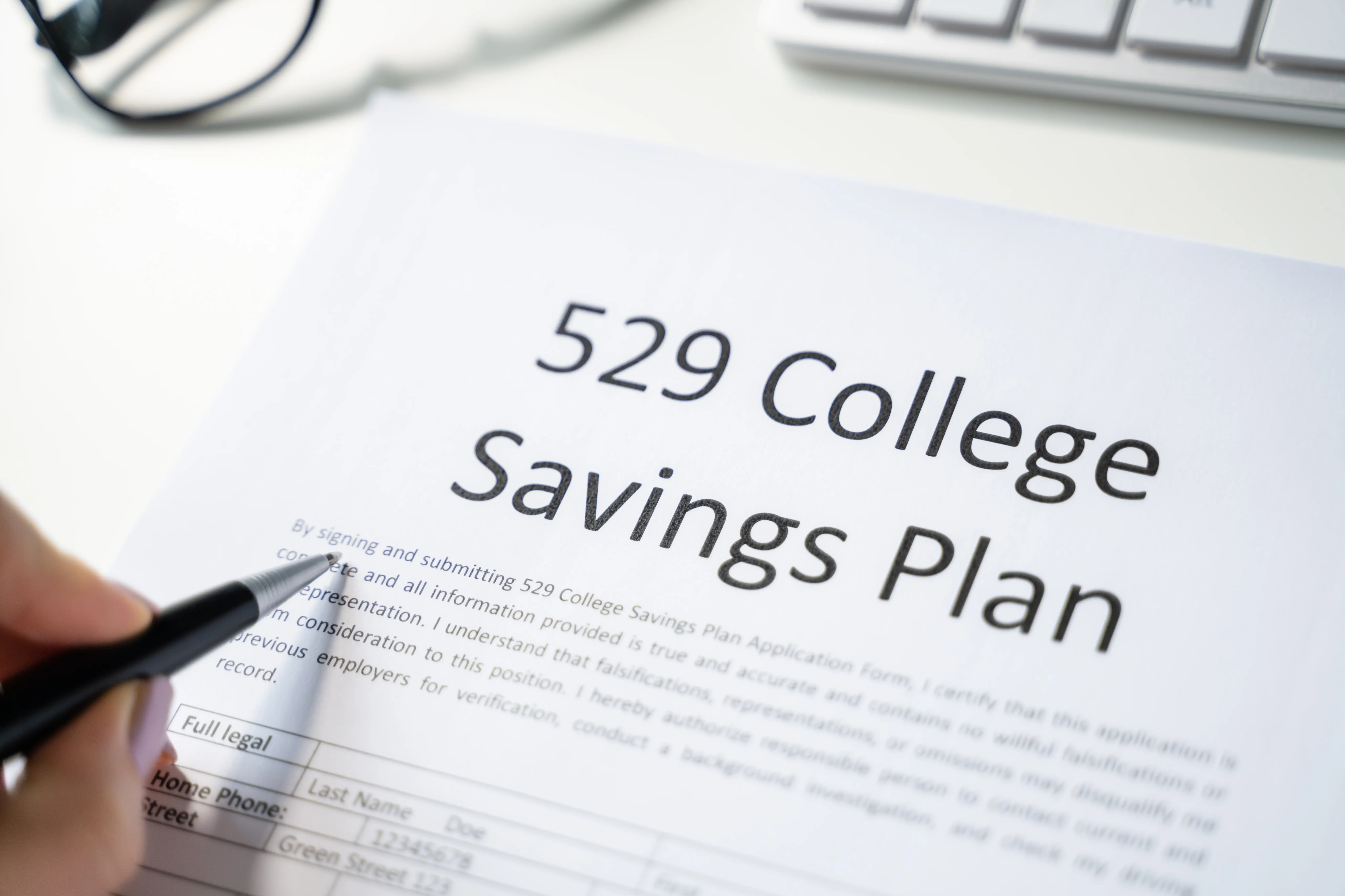 A person holding a pen reviews a document titled "529 College Savings Plan" next to a computer keyboard.