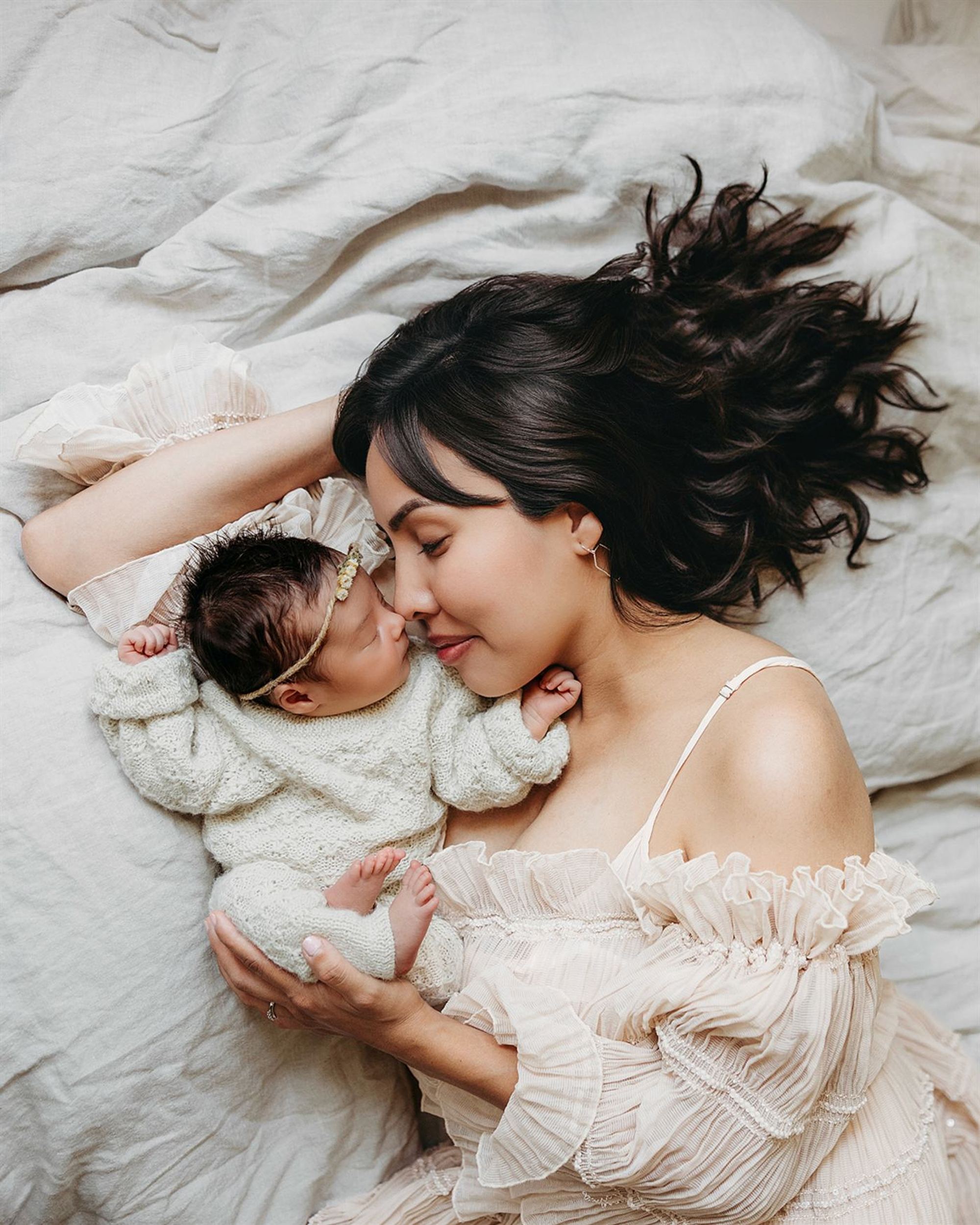 A mother lying in white linen, nose to nose with her sleeping newborn