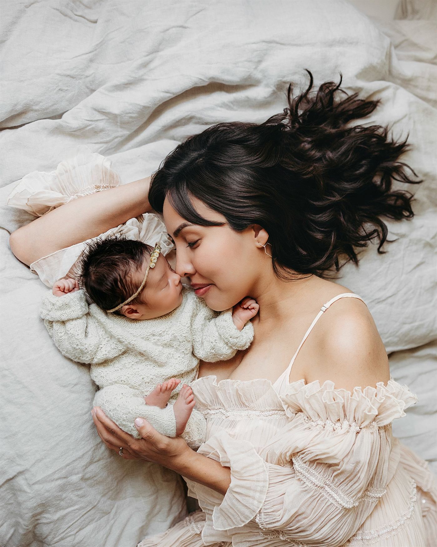 A mother lying in white linen, nose to nose with her sleeping newborn