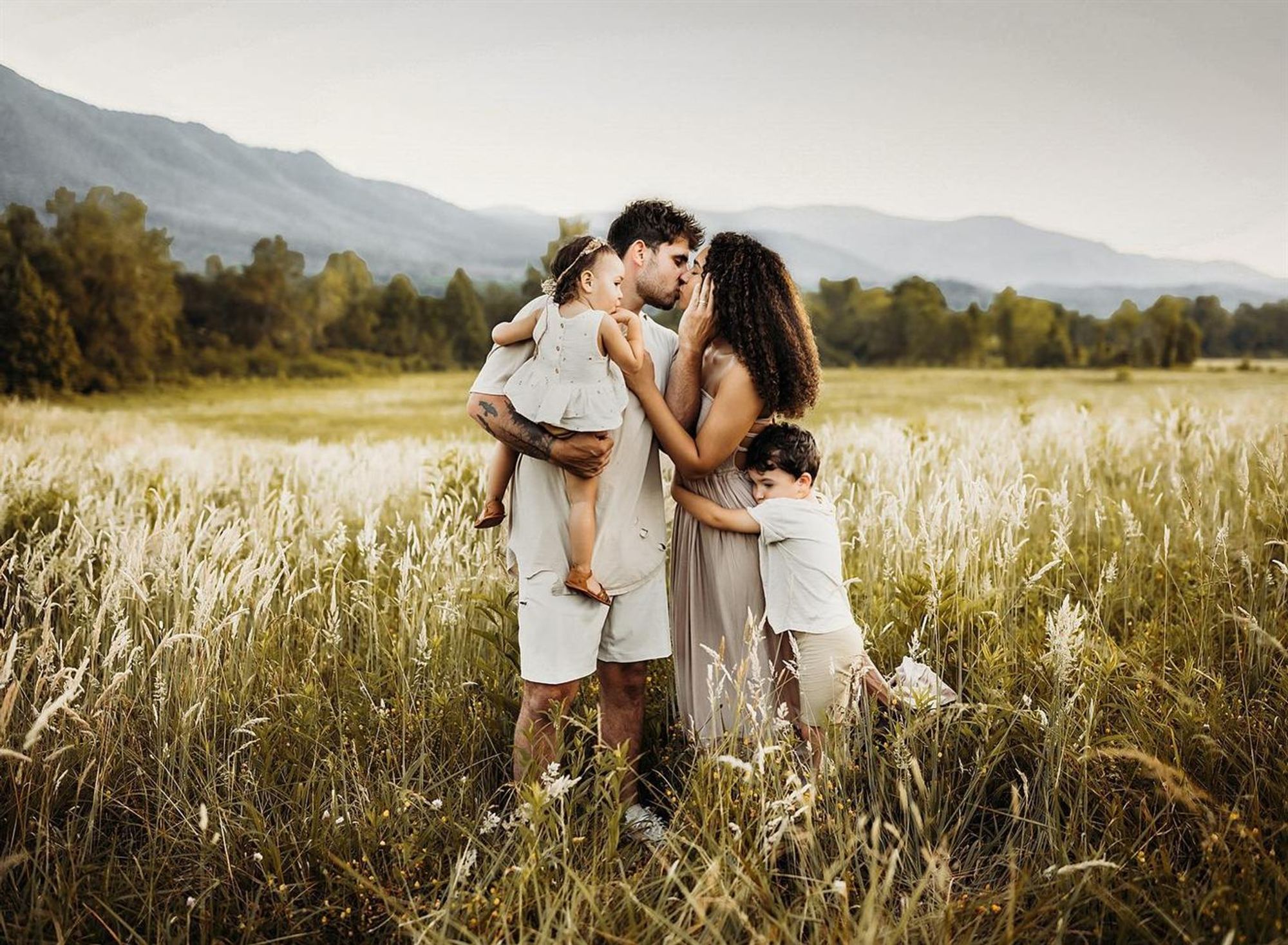 A young family embracing in tall golden grass with mountains behind them