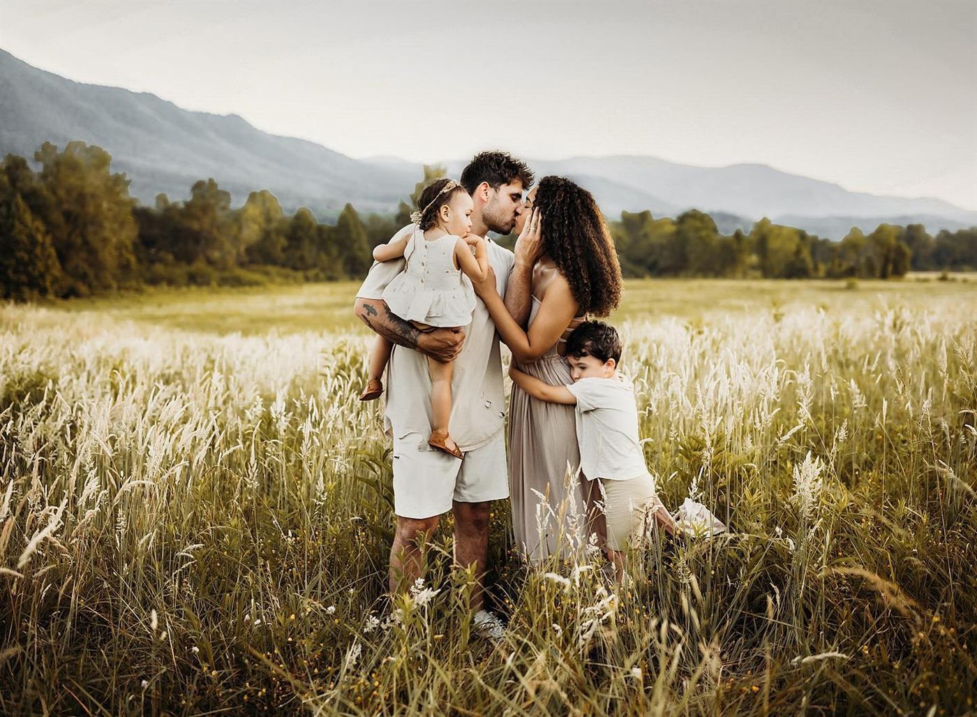 A young family embracing in tall golden grass with mountains behind them