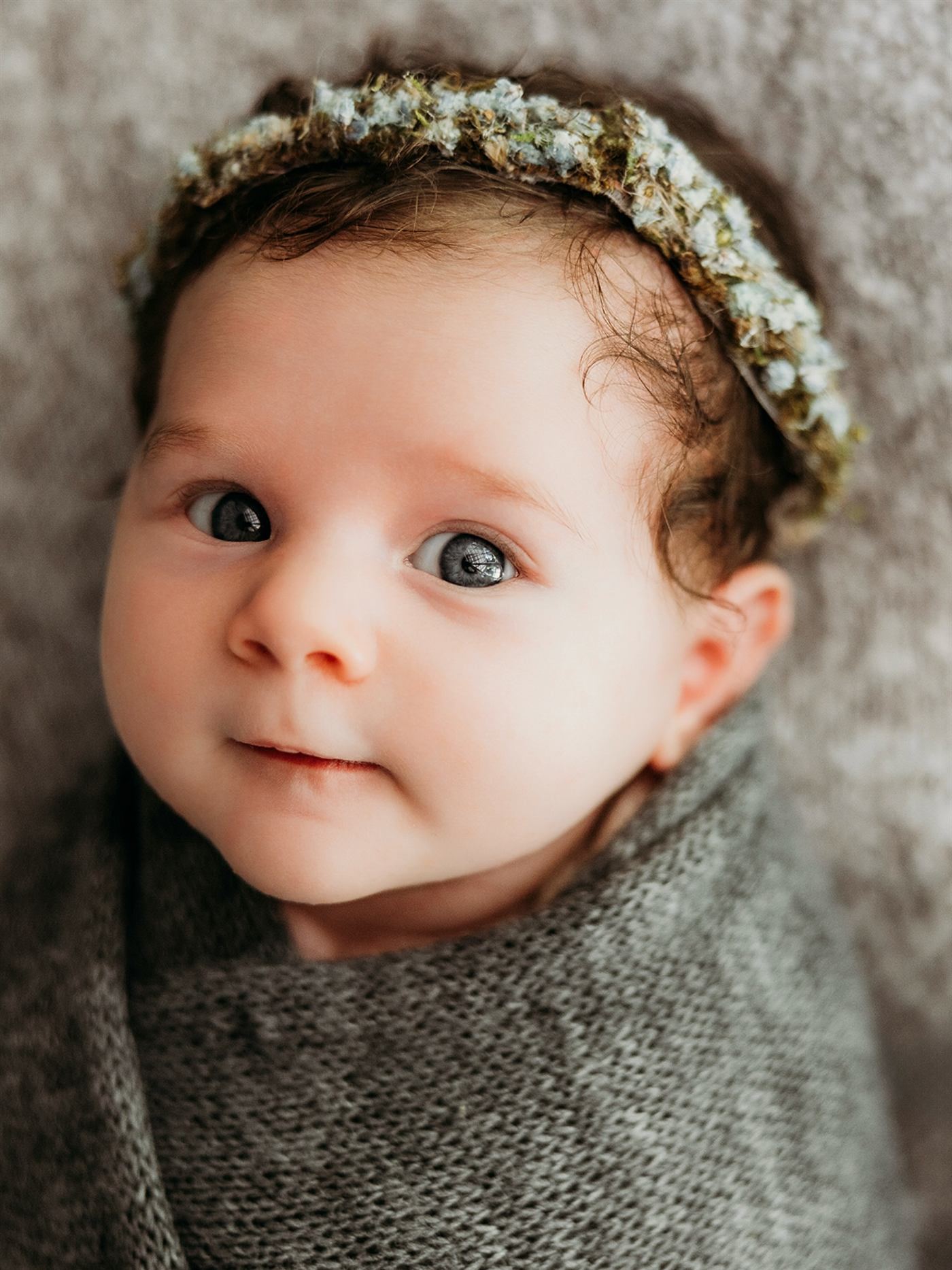 A wide-eyed newborn wrapped in grey knit, wearing a floral halo