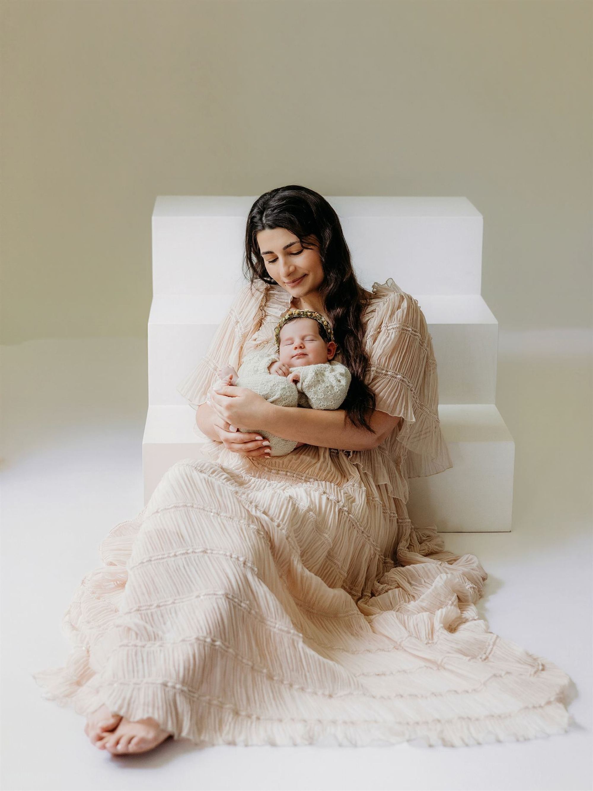 A mother in a cream gown holding her newborn in a softly lit studio