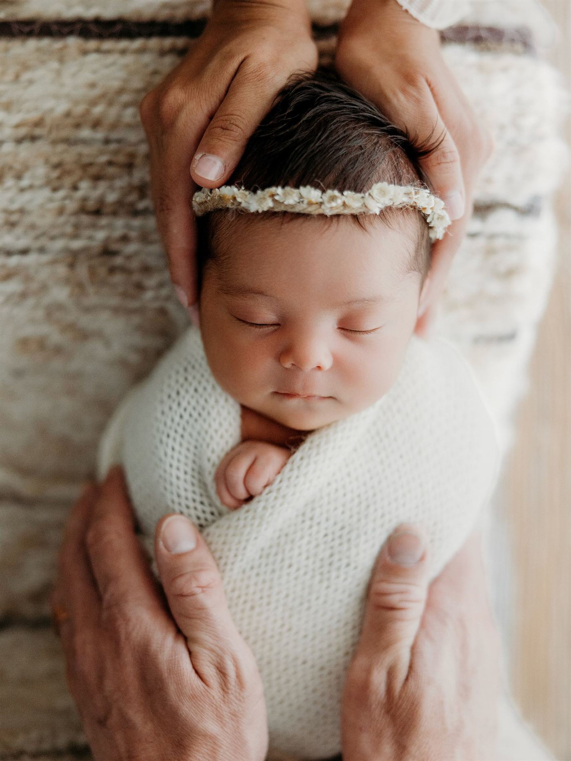 A sleeping newborn with a floral halo, cradled in her parent's hands