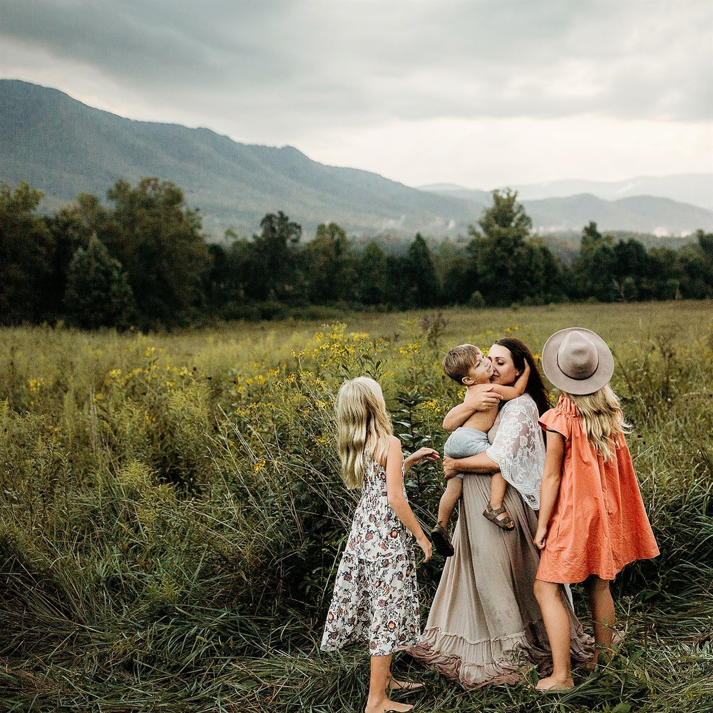 A mother holding her toddler while her daughters play in a meadow below the hills