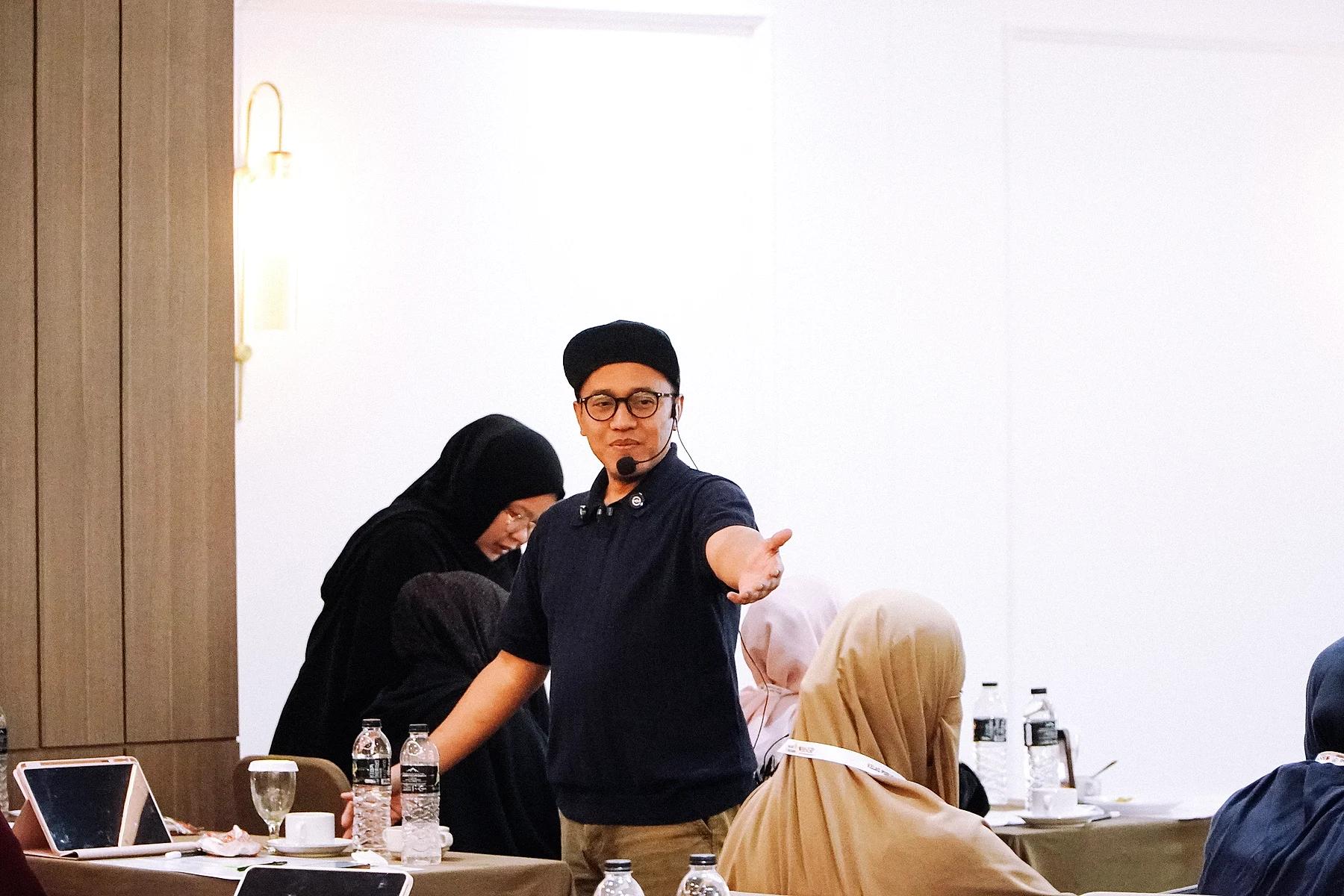 Coach Yoso Lukito memakai headset mikrofon berdiri di antara peserta pelatihan yang duduk di meja dengan tablet dan botol air