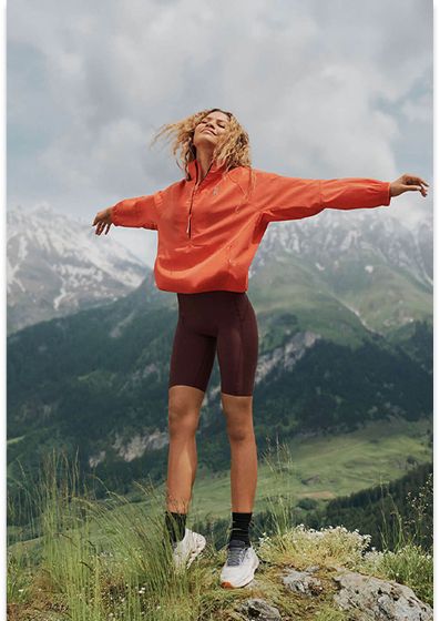 A smiling woman in an orange jacket and dark shorts stands on a mountain peak with arms outstretched, snowy mountains in the background.