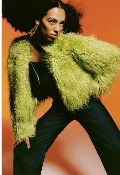 A person with dark curly hair wears a vibrant lime green shaggy jacket and dark jeans, posing against an orange background.