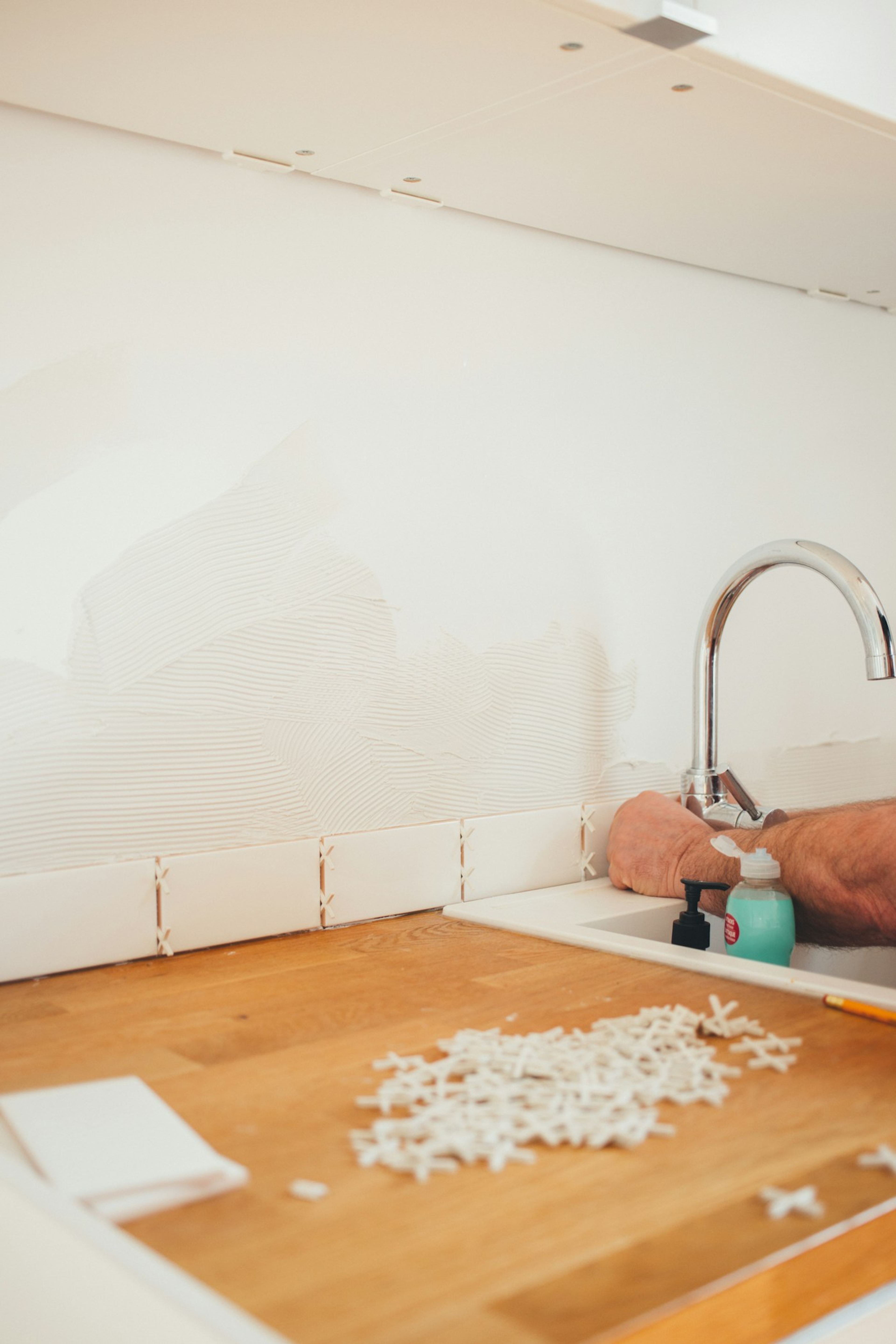 Hands working beside a kitchen tap while a tiled splashback is installed