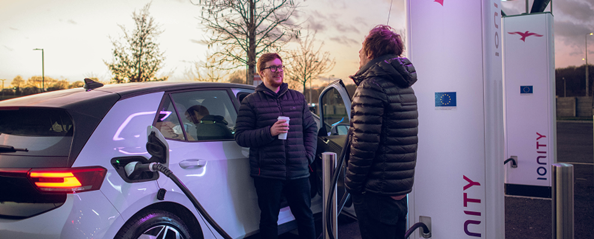 Zwei Männer stehen links neben einem Elektroauto, auf der rechten Seite sieht man Ladeinfrastruktur.