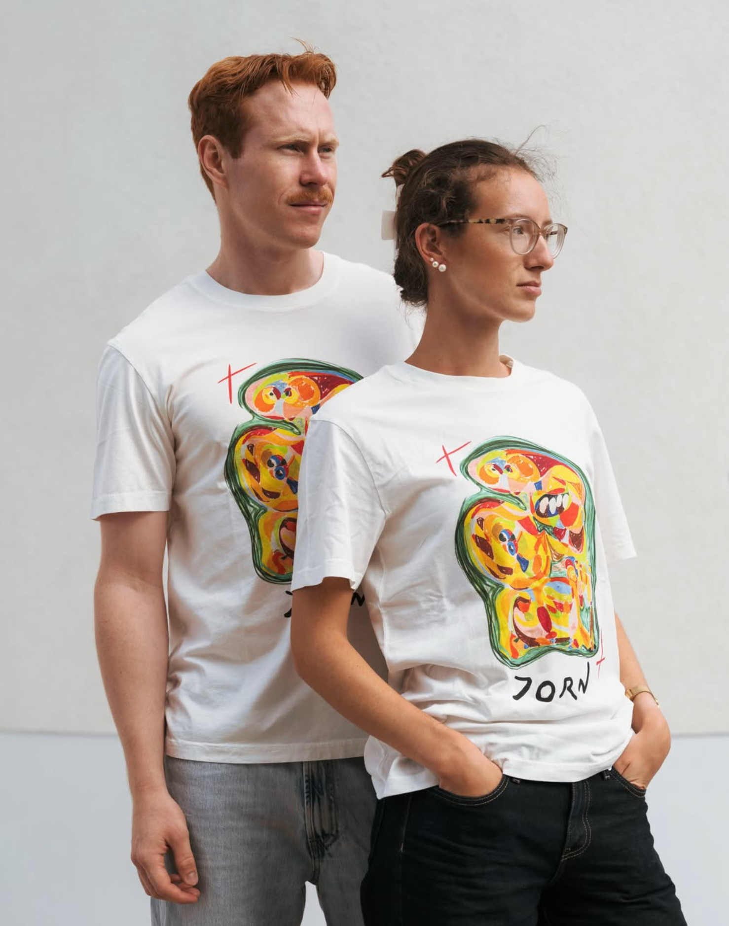 Two models wearing the same white Kunstsilo T-shirt — an Asger Jorn-style expressionist face in yellow, red and green, with the artist’s name below