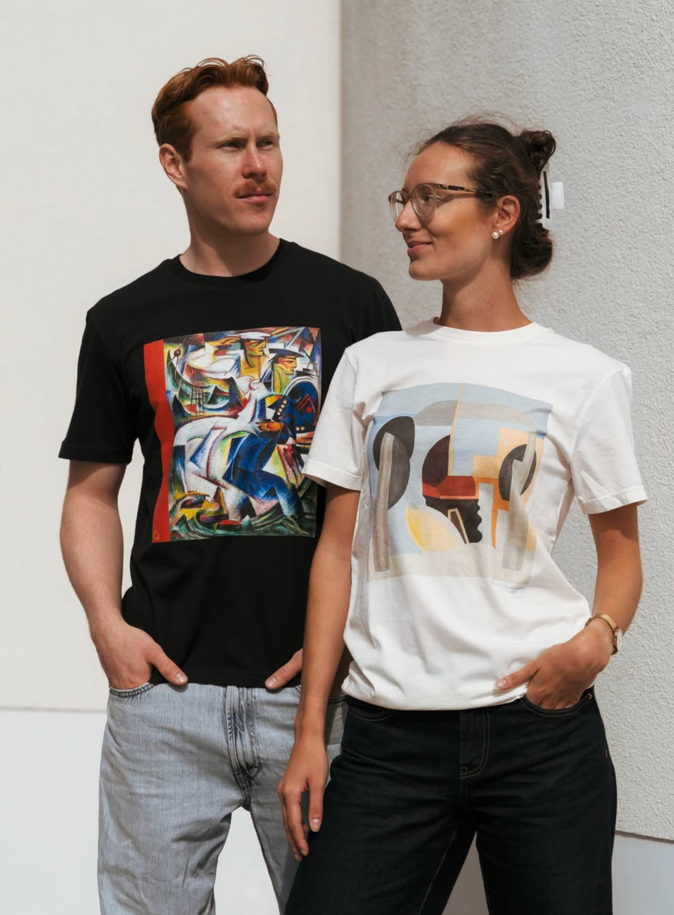 A man and a woman wearing Kunstsilo T-shirts — a black tee with a vivid figurative print and a white tee with an abstract geometric composition, both from the Tangen Collection